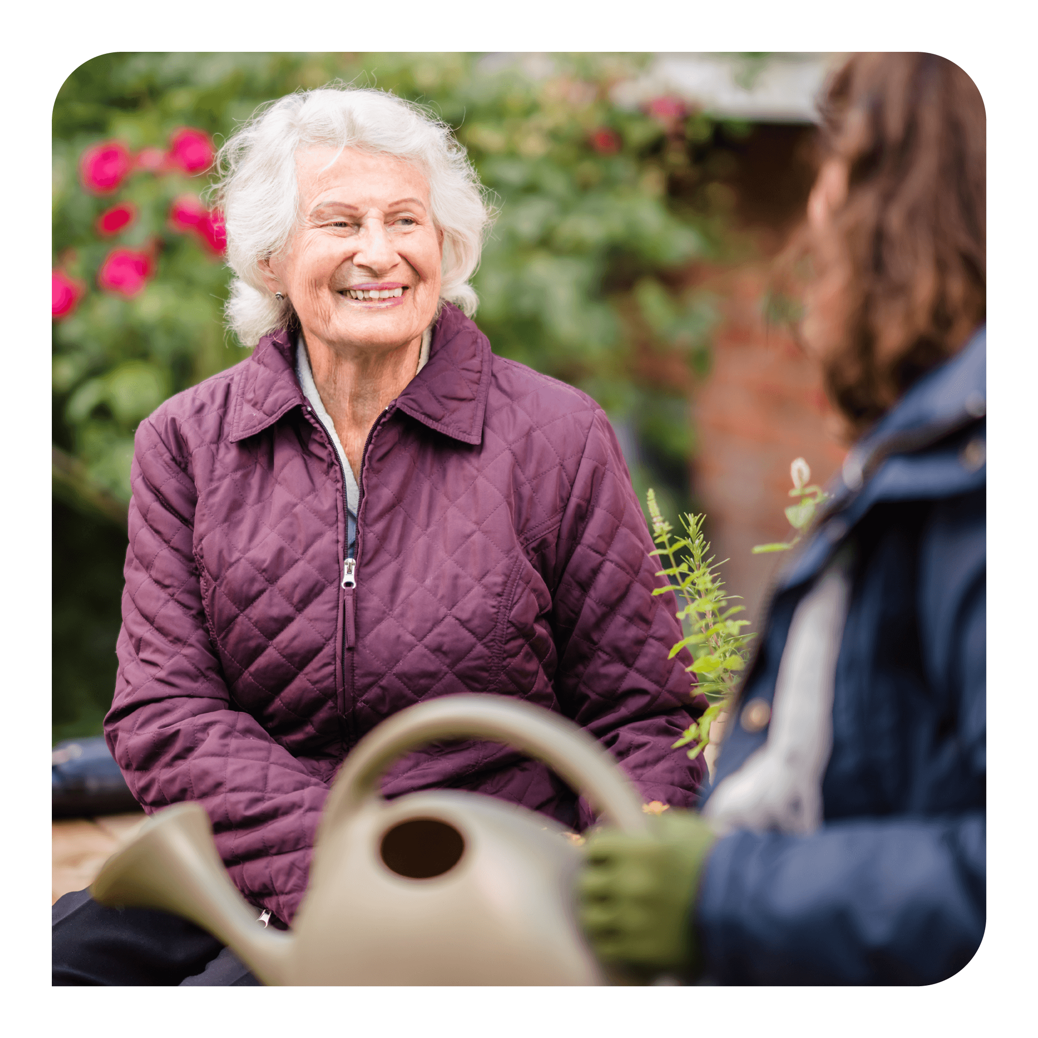 Elderly woman in a purple jacket smiles at another person with a watering can in a garden setting. - Home Instead