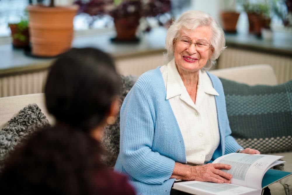 Elderly woman with glasses reading book while Care Professional talks to her - Home Instead