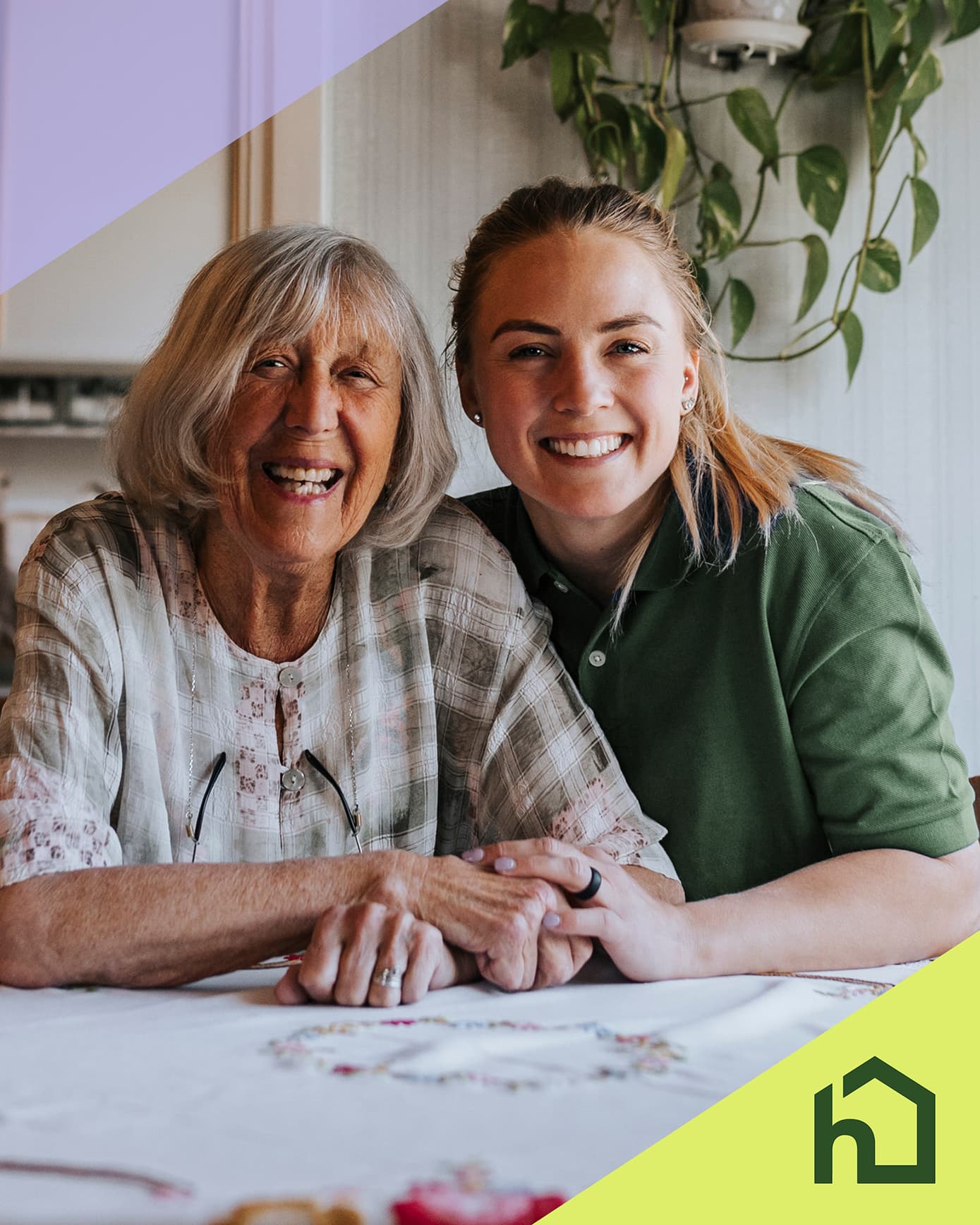 Smiling elderly woman and young caregiver sit together at a table, hands joined, with a house logo in the corner. - Home Instead