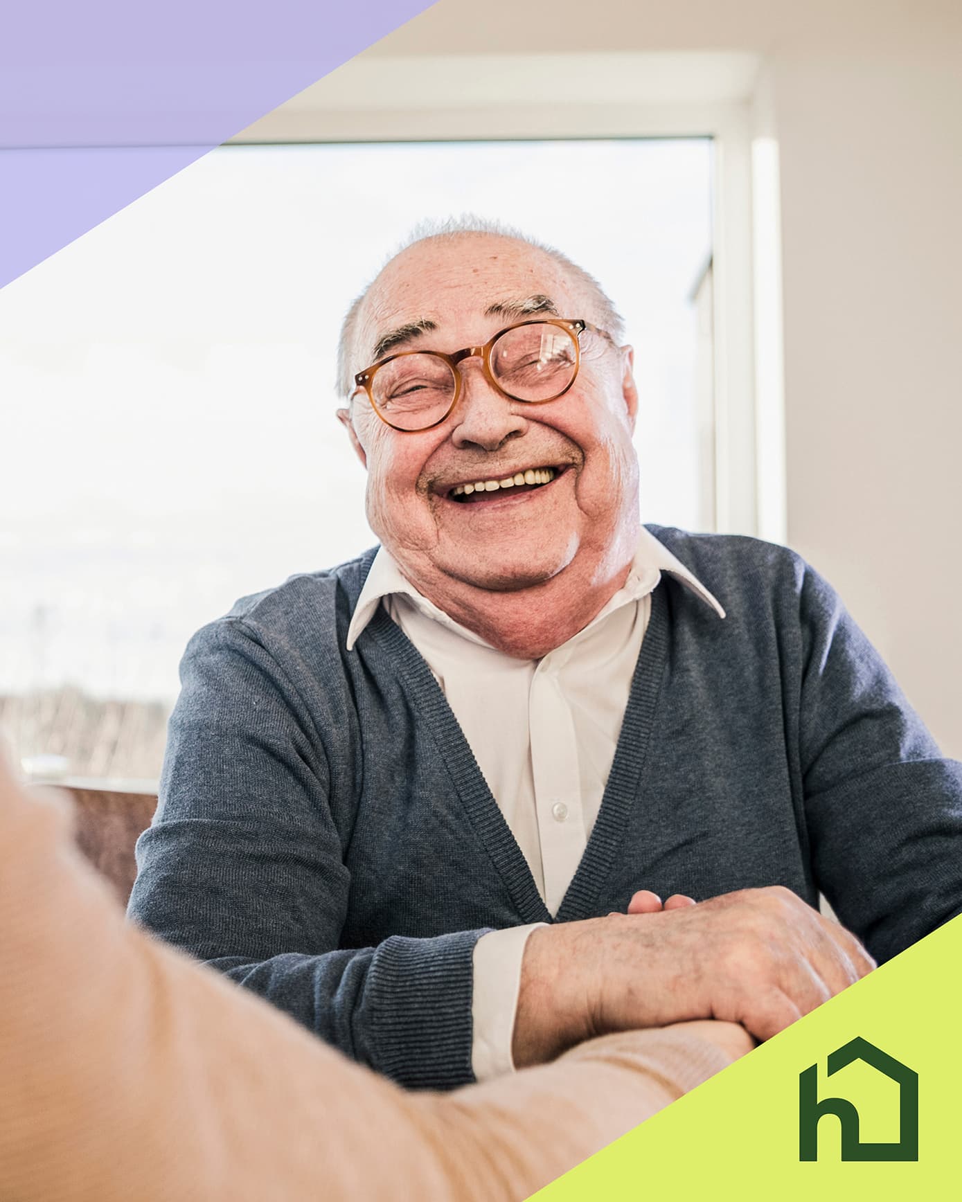 Smiling elderly man in glasses sits indoors, wearing a white shirt and blue sweater, with a house logo in corner. - Home Instead