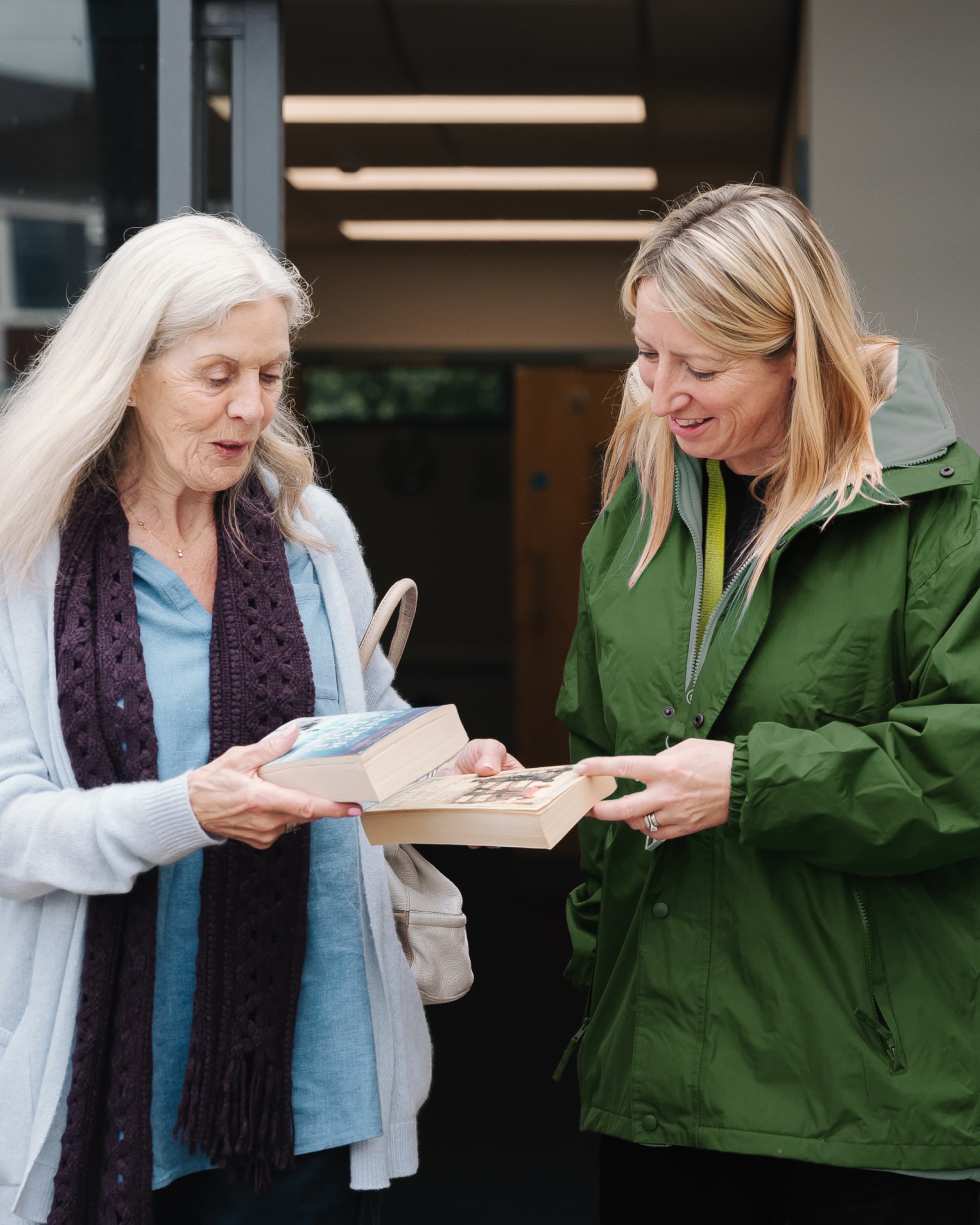 Two women standing together and exchanging books, one wearing a green jacket, the other a purple scarf. - Home Instead