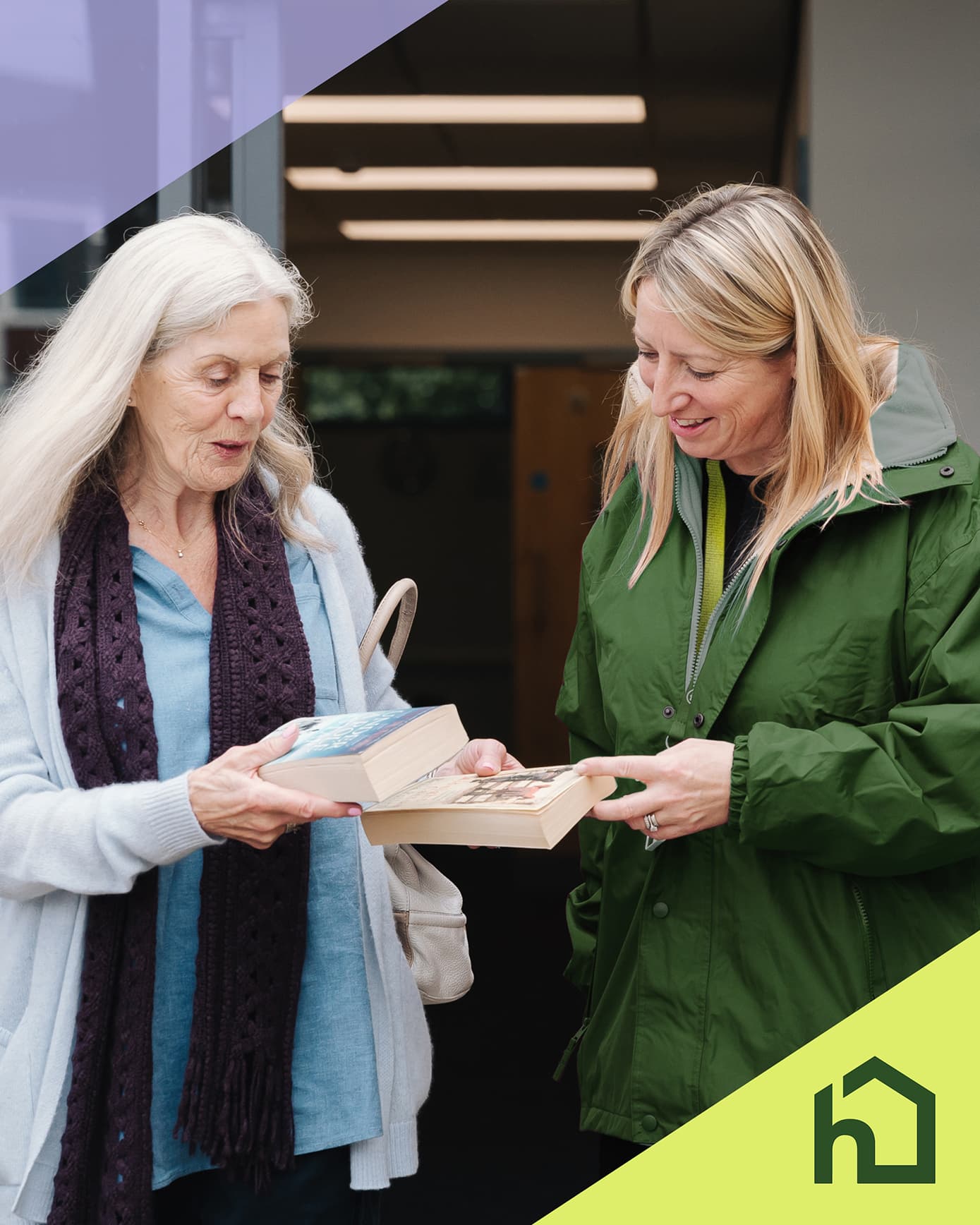 Two women smiling and exchanging books indoors, with a green logo in the bottom right corner. - Home Instead