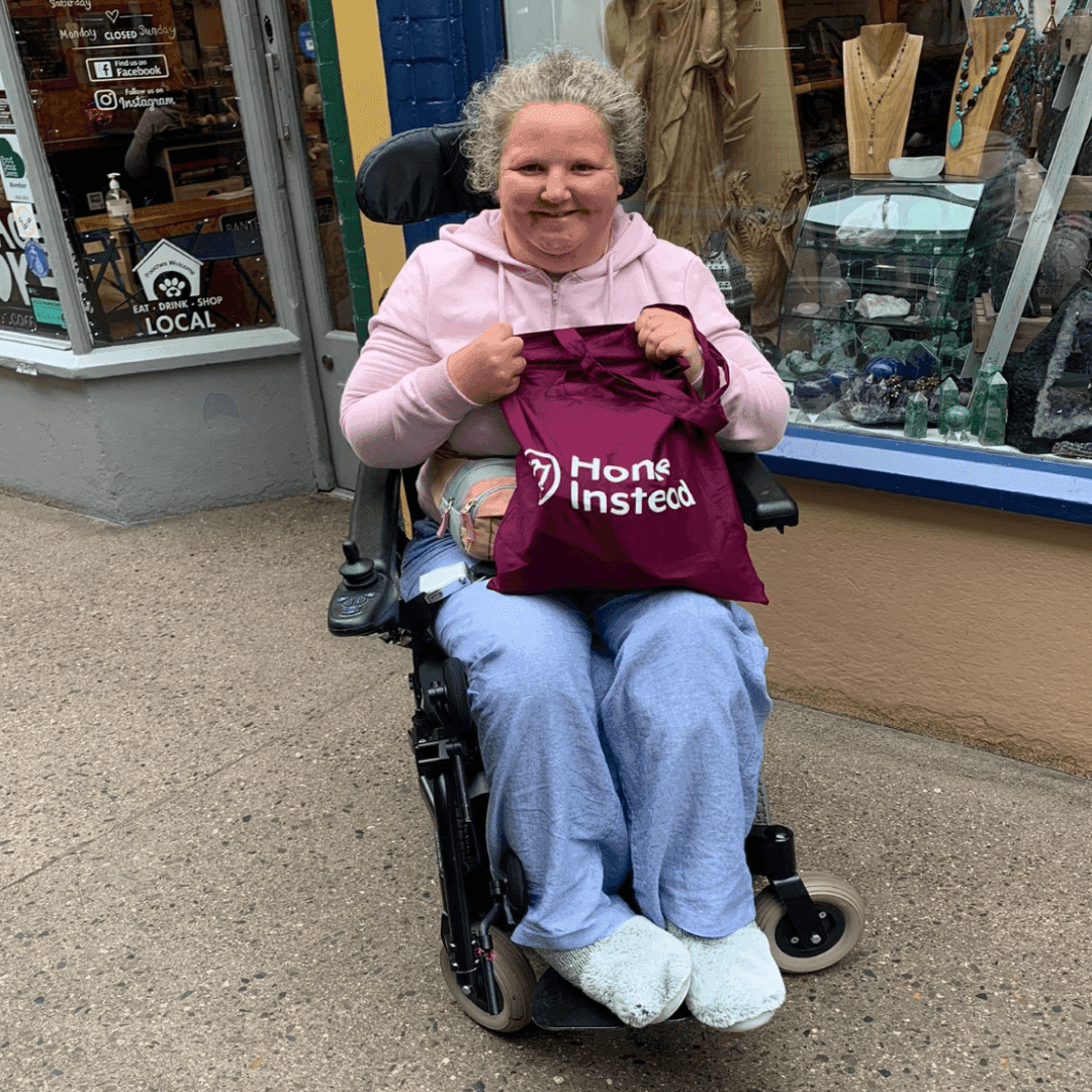 Person sitting in a wheelchair holding a purple Home Instead bag, smiling outside a store with colorful window displays. - Home Instead