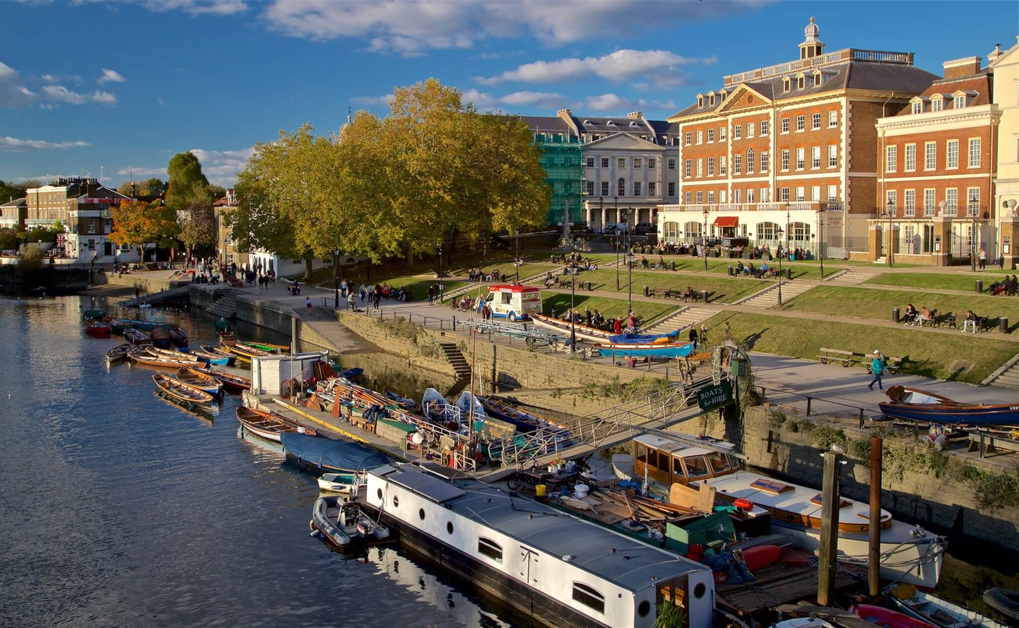People relaxing on grassy steps by the riverside, with boats docked in the water and buildings in the background. - Home Instead