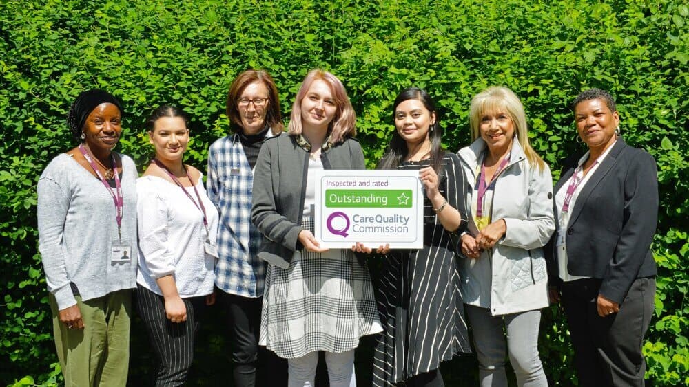 A diverse group of smiling individuals holding a Care Quality Commission sign marked "Outstanding," standing outdoors. - Home Instead