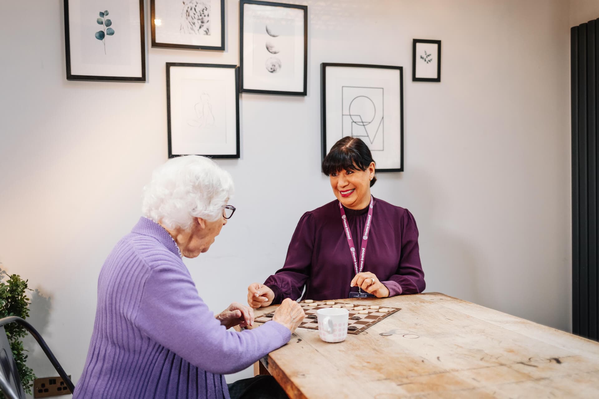 Two women sit at a table playing a board game in a room with framed artwork on the walls. - Home Instead