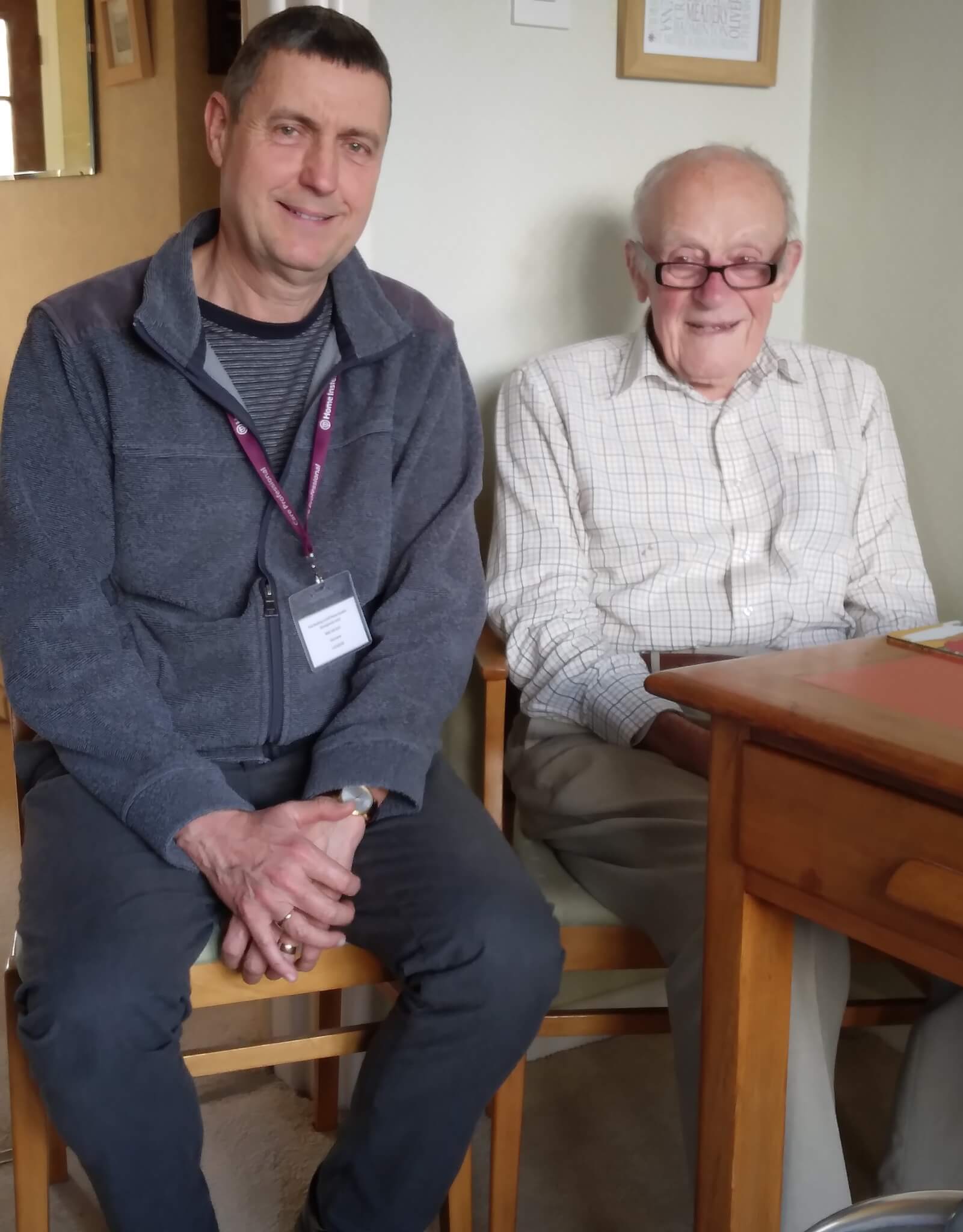 Two men sitting side by side indoors, one wearing a name badge and the other in a checkered shirt, both smiling. - Home Instead