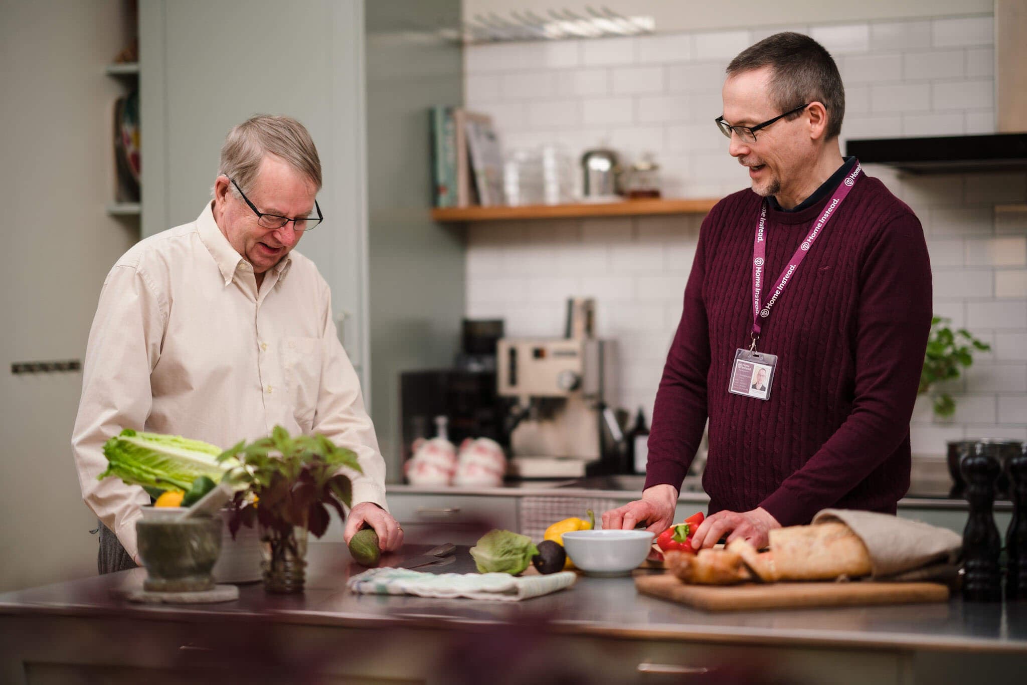 Two men smile while preparing vegetables together in a kitchen with various ingredients on the counter. - Home Instead
