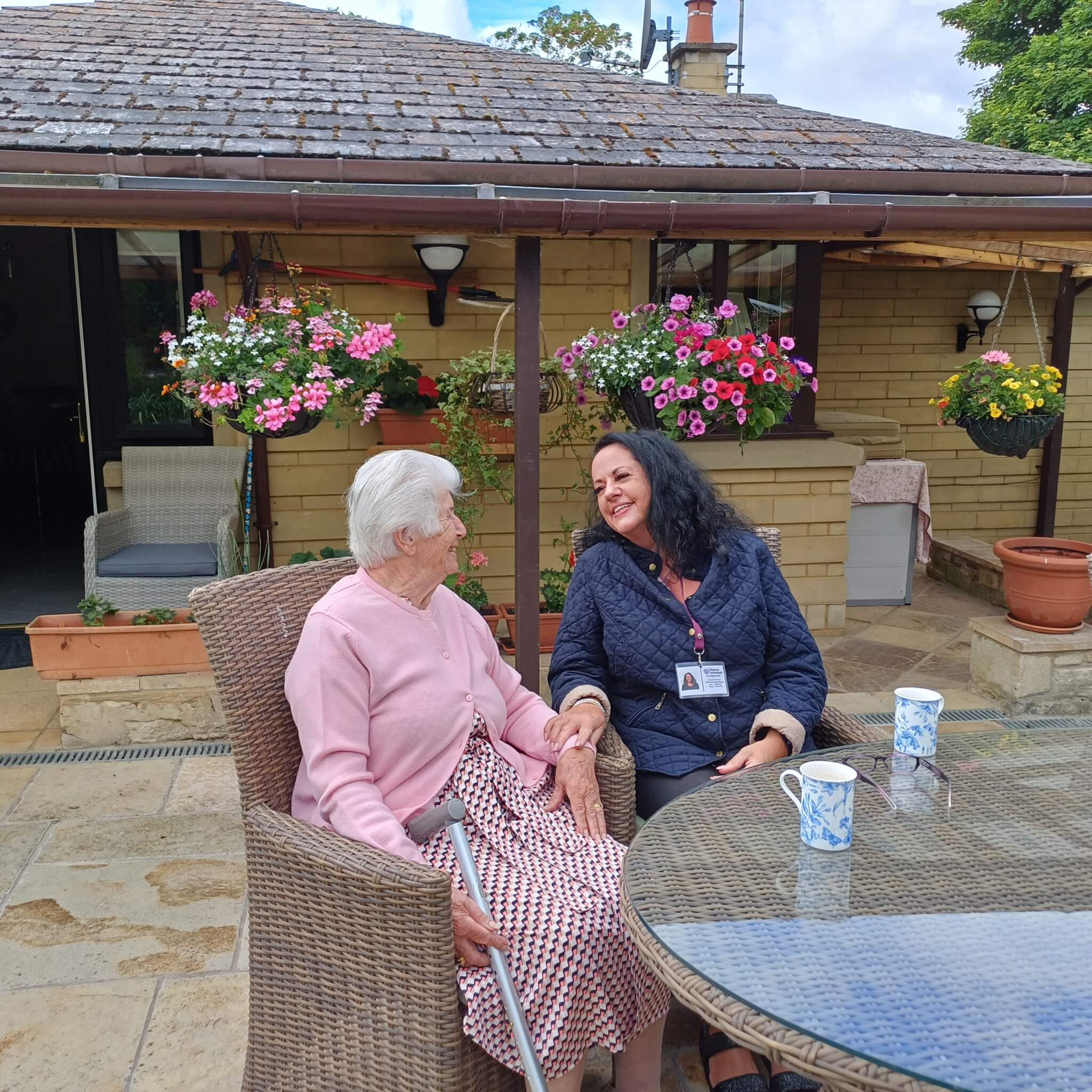 An elderly woman and a caregiver share smiles at a patio table outside with flowers hanging nearby. - Home Instead