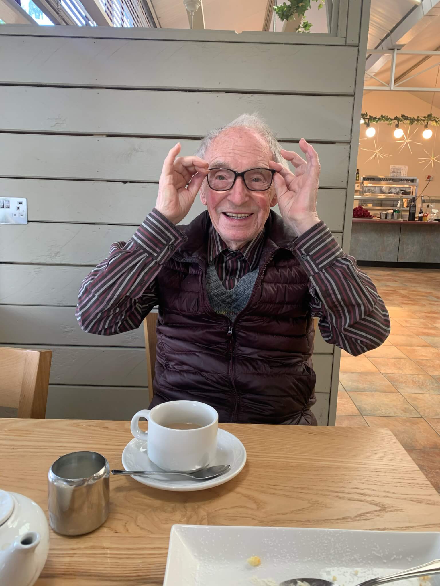 An elderly man wearing glasses smiles, sitting at a table with a cup of tea in a cozy café. - Home Instead