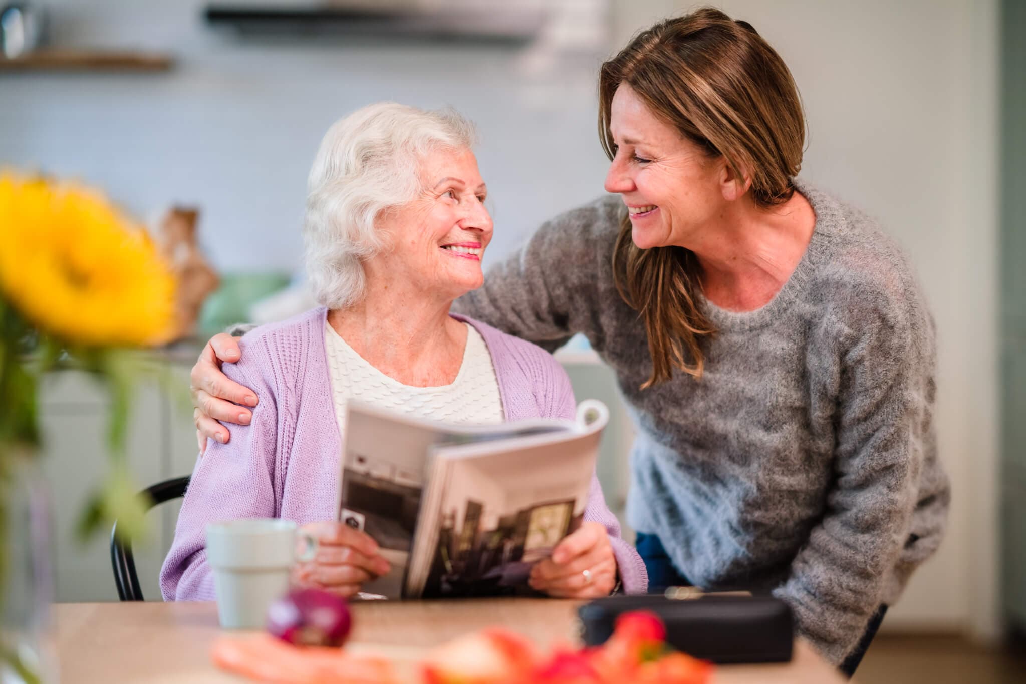 Older woman reading a magazine, smiling at a younger woman embracing her. Sunflower and fruits in the foreground. - Home Instead