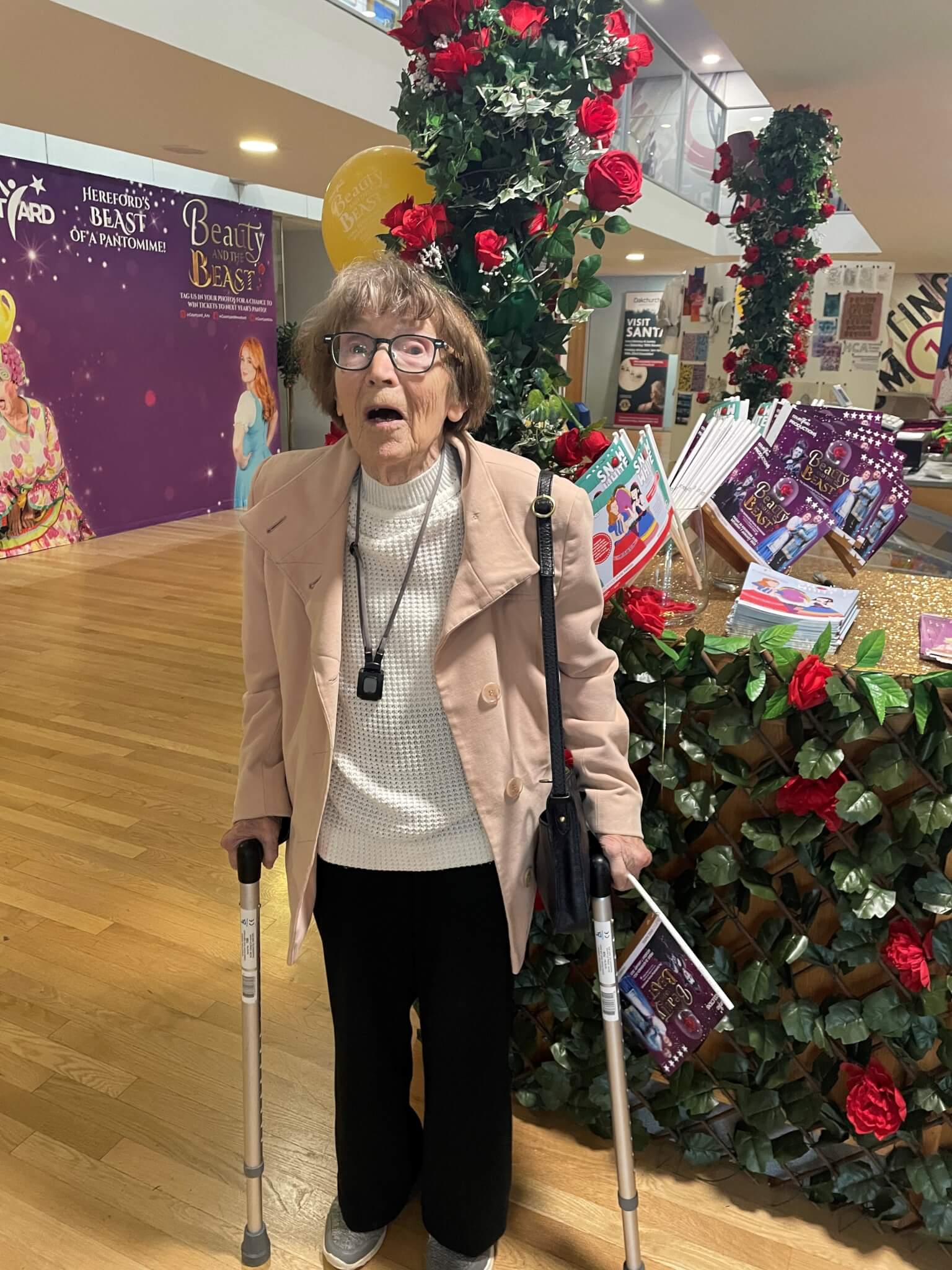 Elderly woman with a surprised expression, using crutches, standing near a decorated display for "Beauty and the Beast". - Home Instead