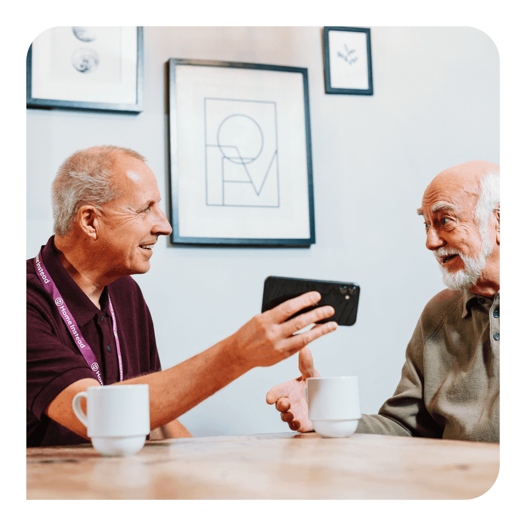 Two elderly men smiling and engaging in conversation, holding a phone, with coffee mugs on the table. - Home Instead
