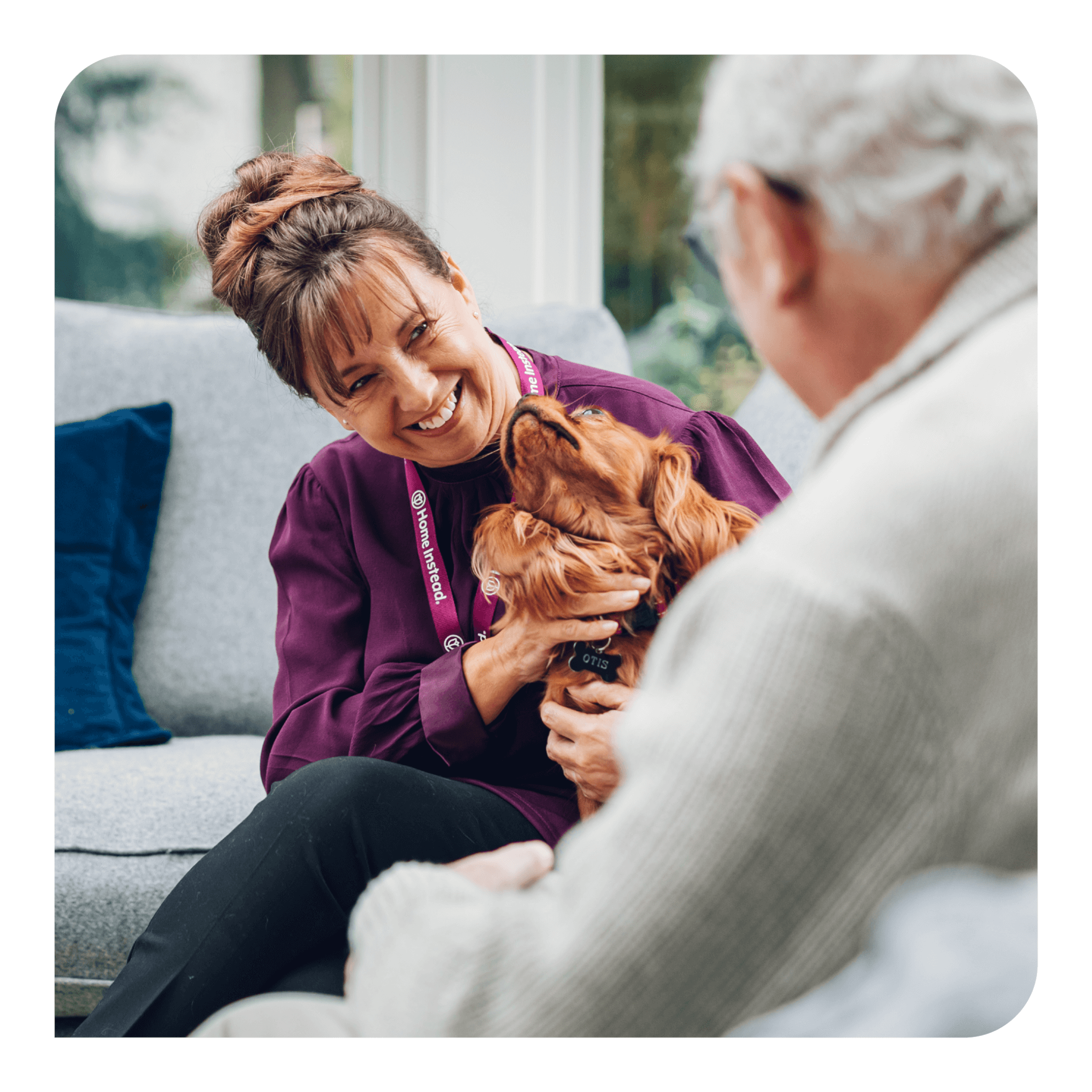 A woman smiles at an elderly man while petting a brown dog. They are seated in a cozy living room setting. - Home Instead