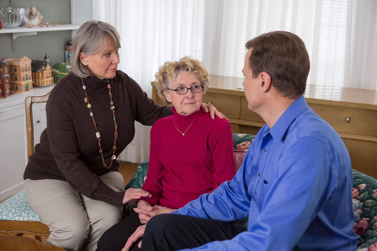 Three people having a serious conversation in a living room: an elderly woman and two adults, with one comforting her. - Home Instead