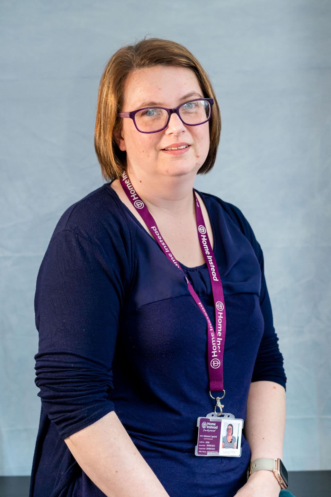A woman with glasses, wearing a blue shirt and a purple lanyard with an ID badge, poses in front of a plain background. - Home Instead