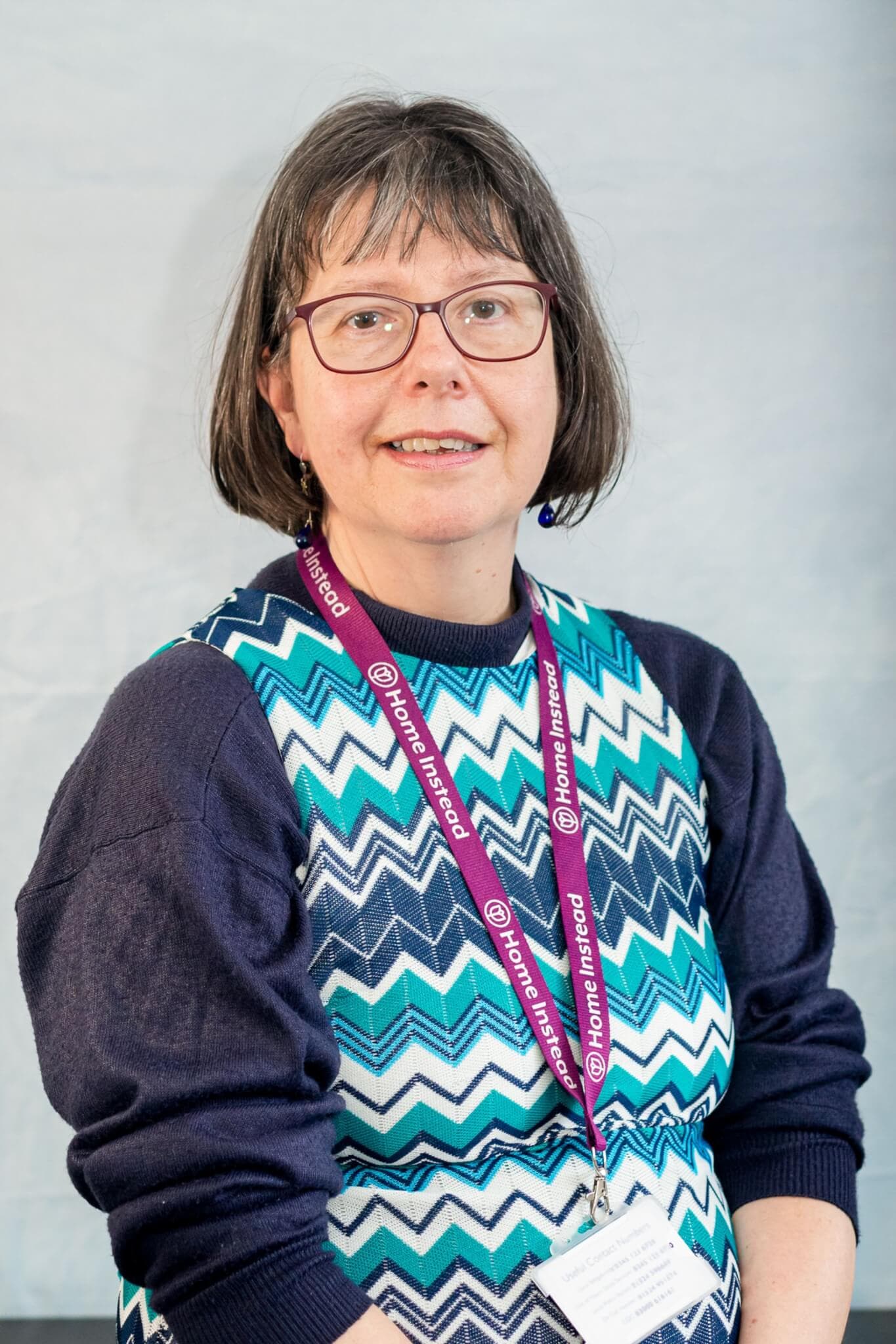 A woman wearing glasses and a patterned dress with a "Home Instead" lanyard smiles at the camera. - Home Instead