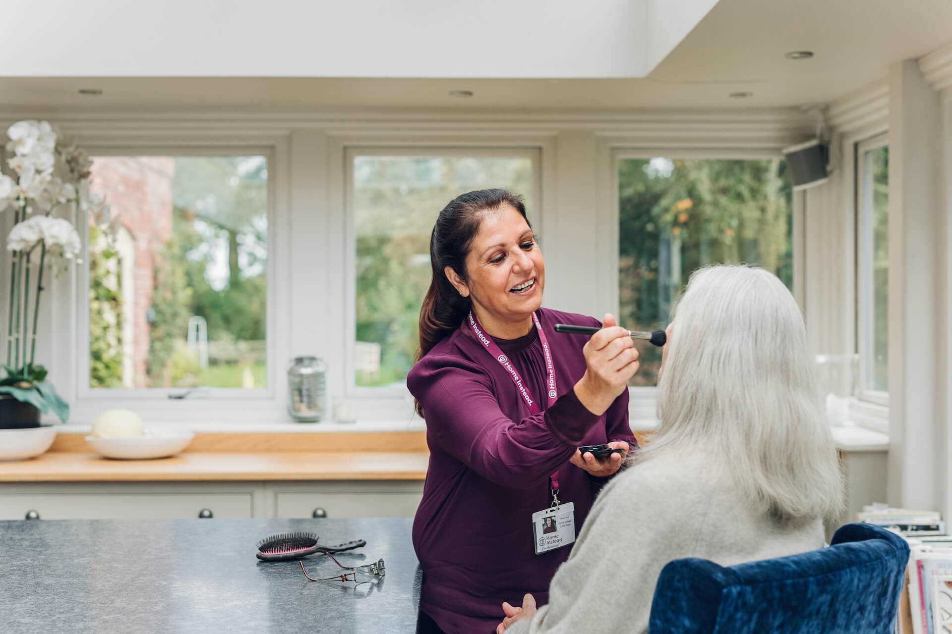 A woman in a maroon uniform applying makeup to a seated elderly woman in a bright room. - Home Instead