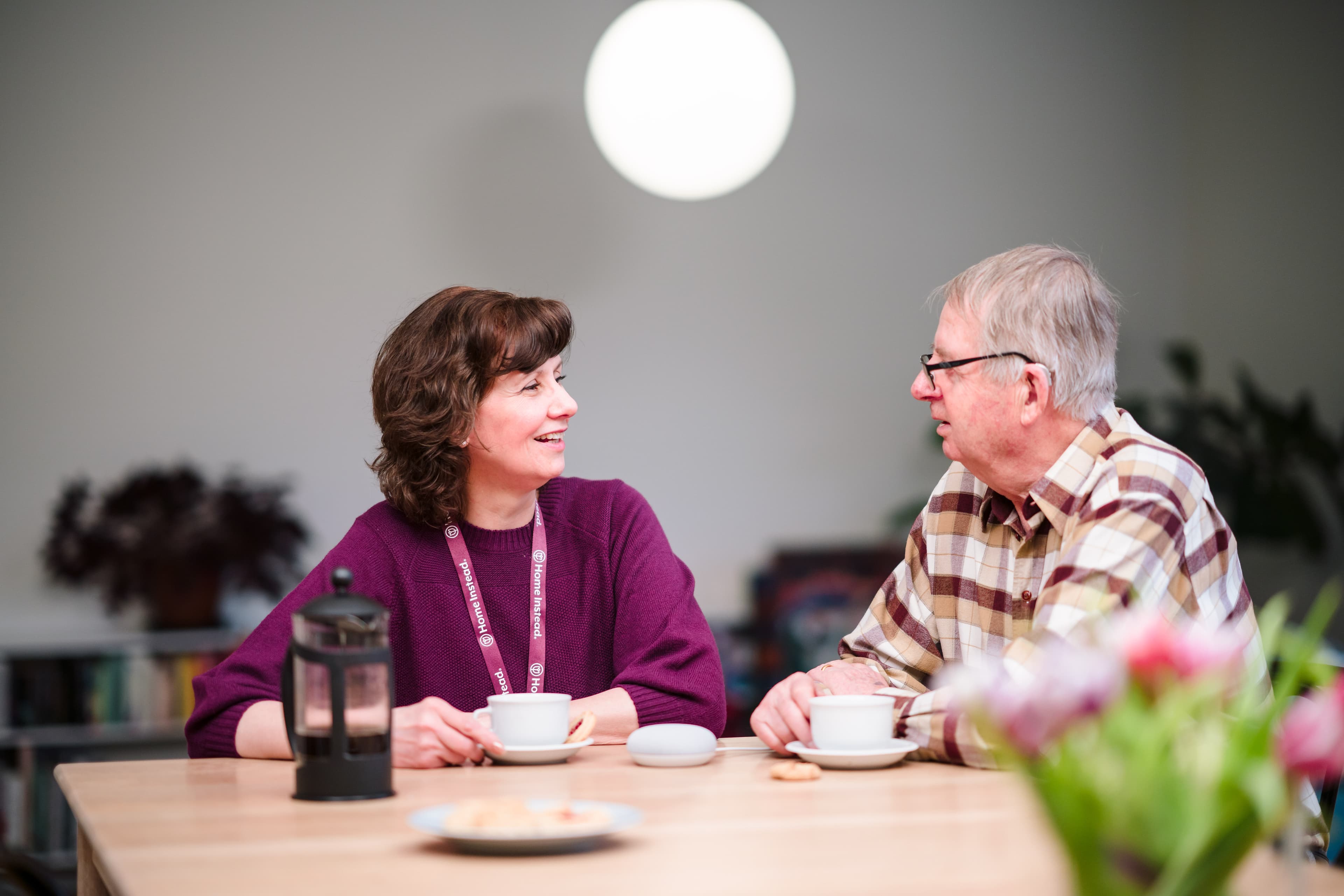 Two people sit at a table with coffee, engaging in conversation. A plant and flowers are in the background. - Home Instead