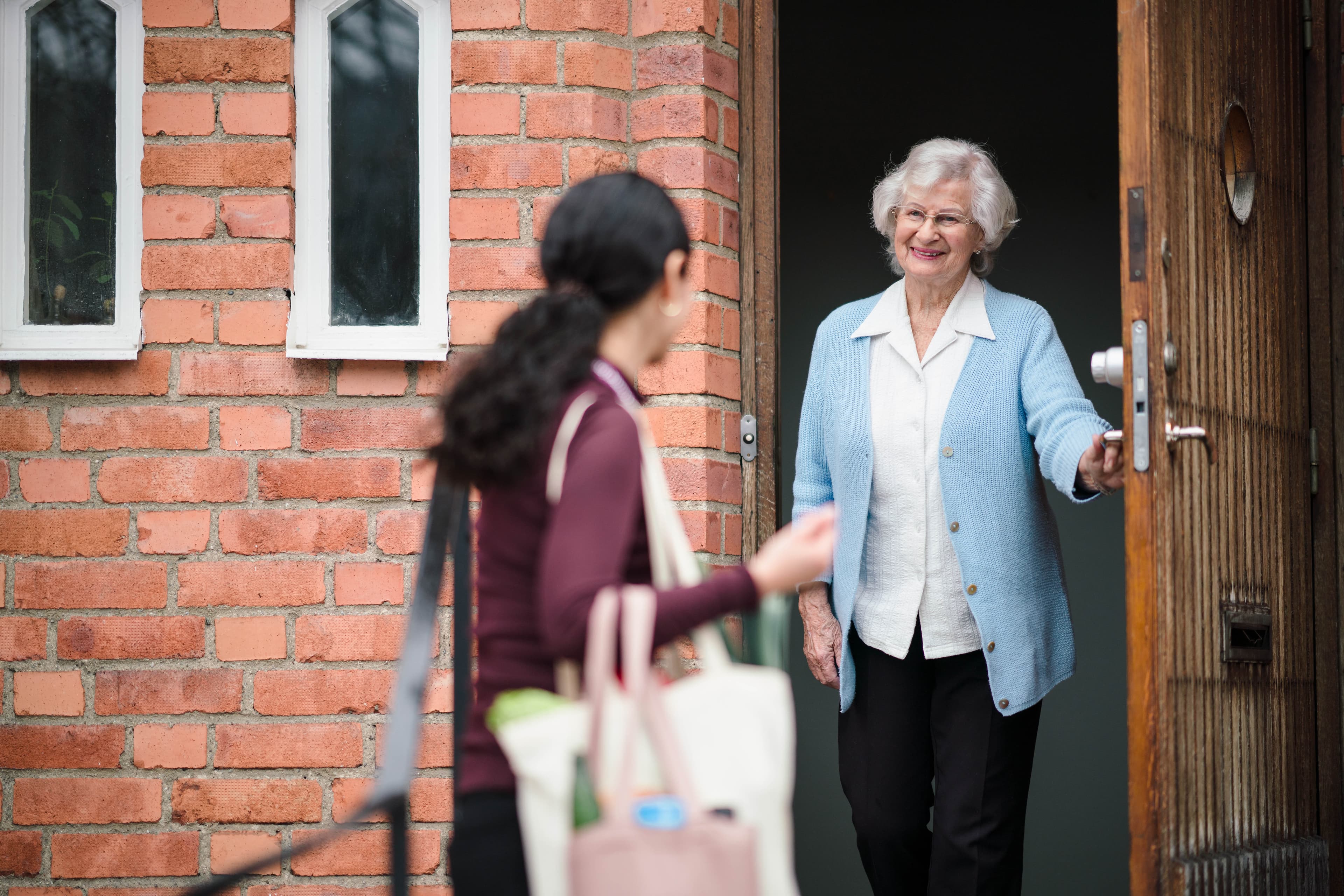 An elderly woman with white hair opens the door to greet a visitor carrying bags, against a brick wall background. - Home Instead