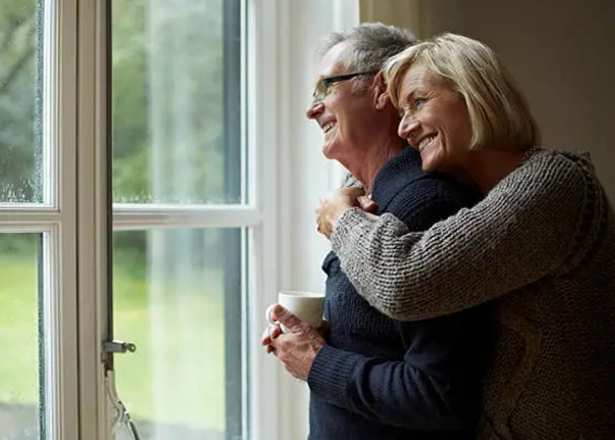 A smiling elderly couple embraces while looking out a window, with light streaming in and greenery visible outside. - Home Instead