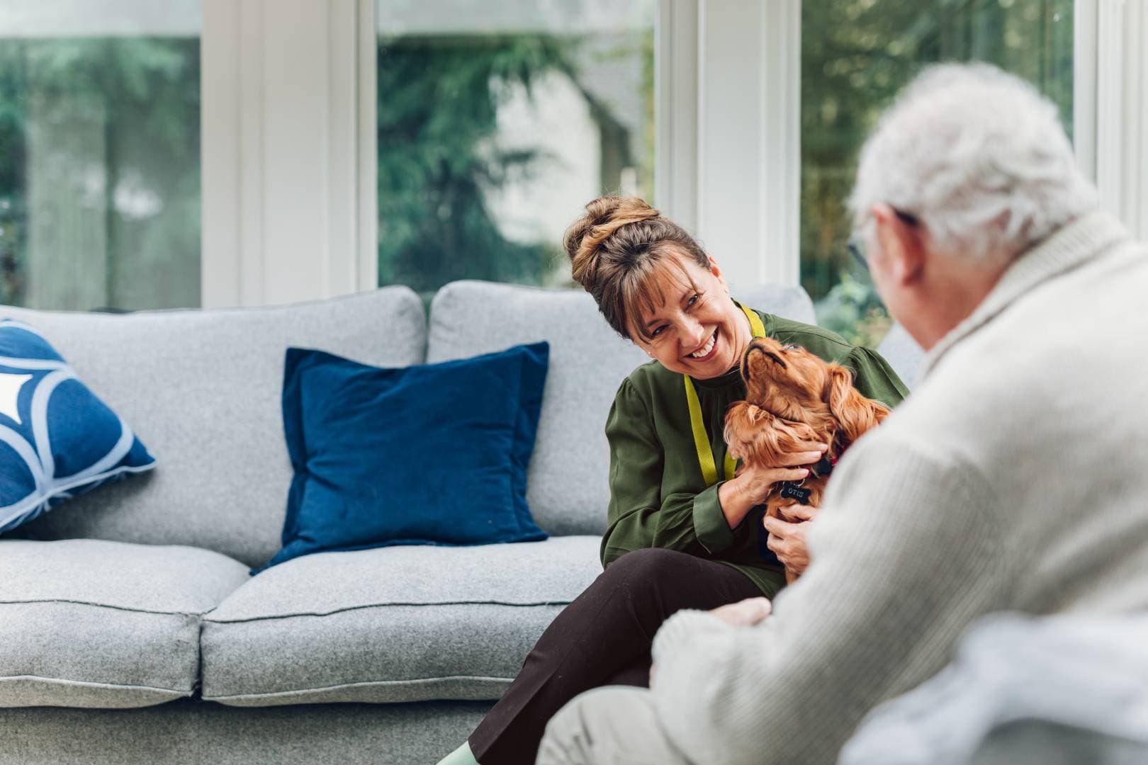 Woman smiles holding a dog on a sofa while an older man sits nearby in a bright lounge. - Home Instead