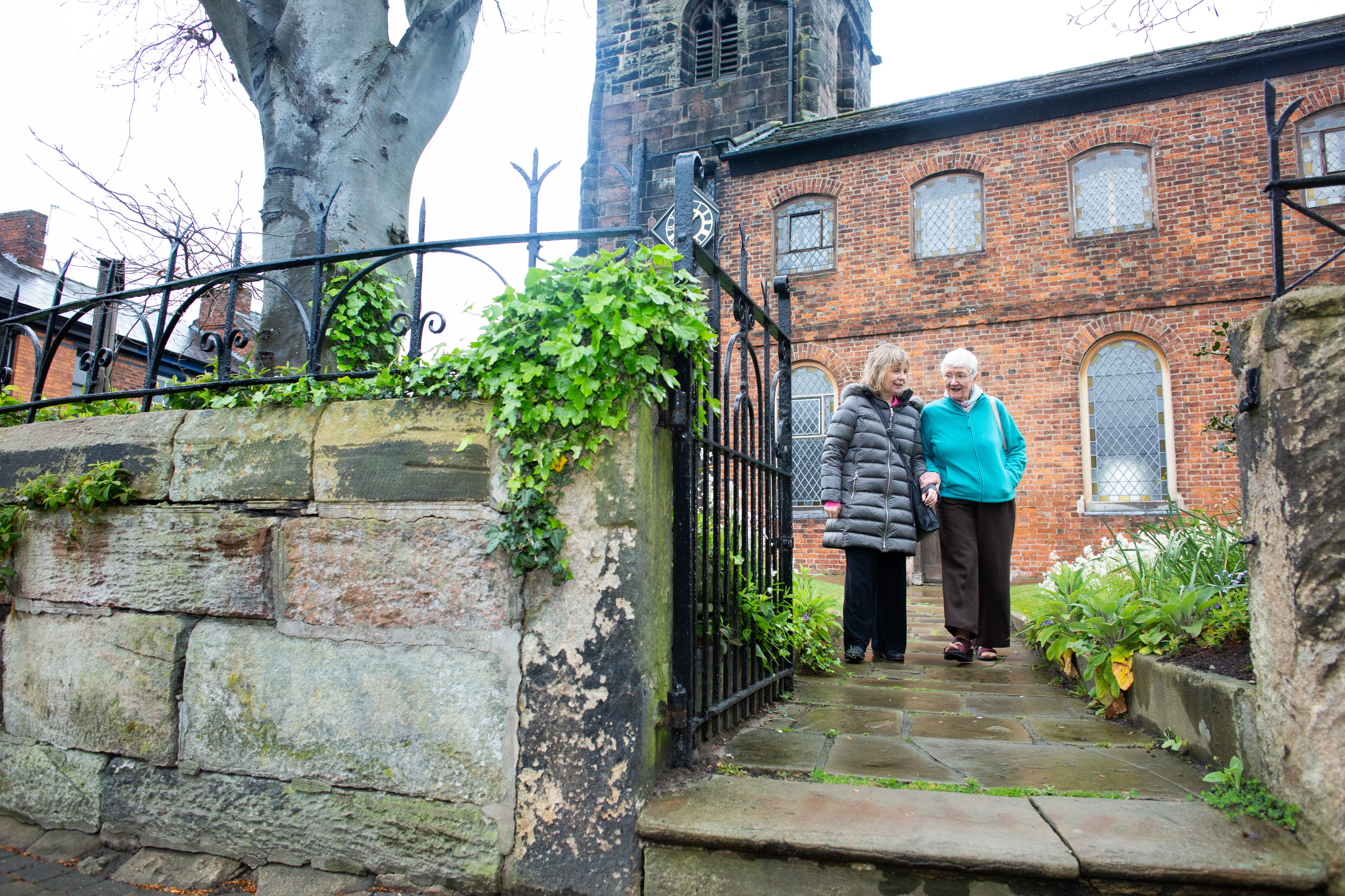 Two elderly women holding hands and walking down a stone path by a brick building with iron fencing and ivy. - Home Instead