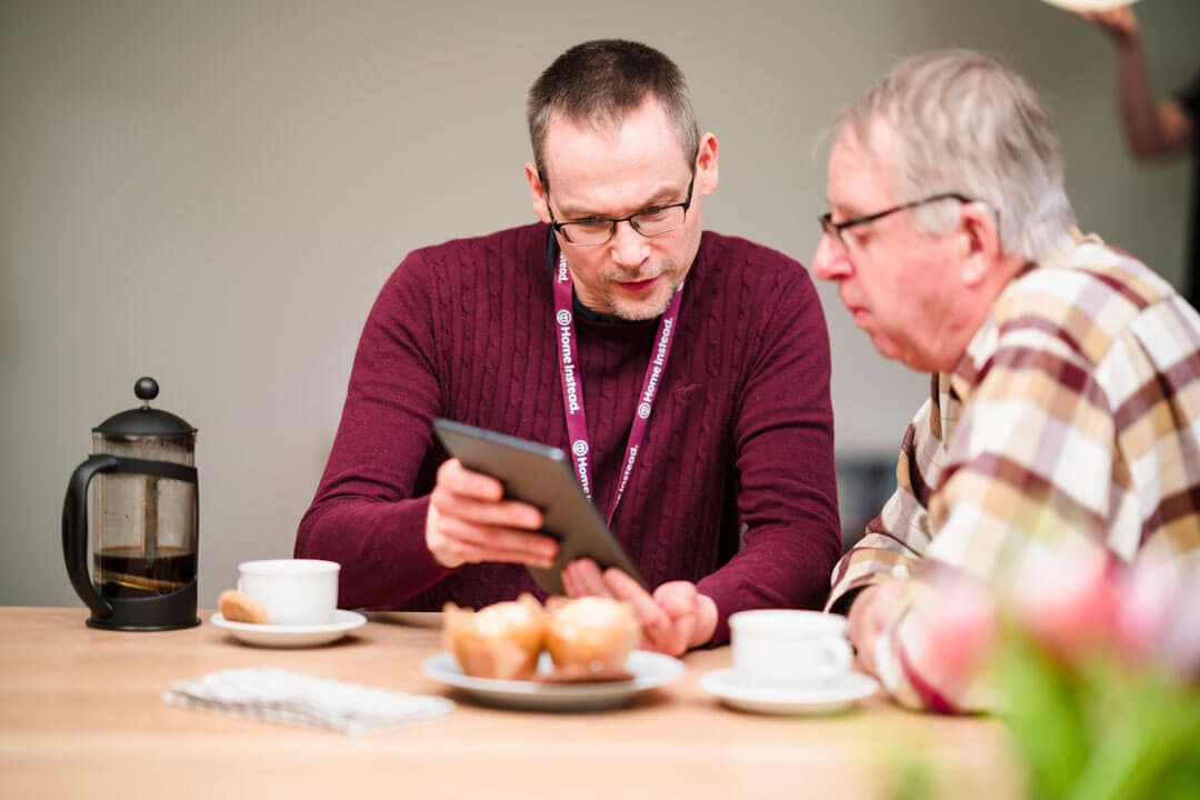 Two older adults looking at a tablet together at a table with pastries, a coffee pot, and cups. - Home Instead