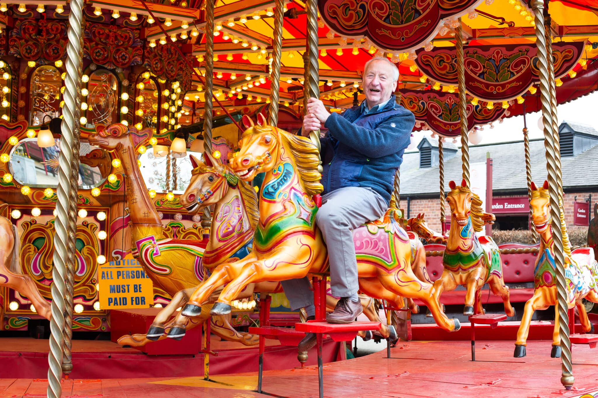 An older man in a blue jacket rides a colorful carousel horse, smiling and holding the pole, with buildings in the background. - Home Instead