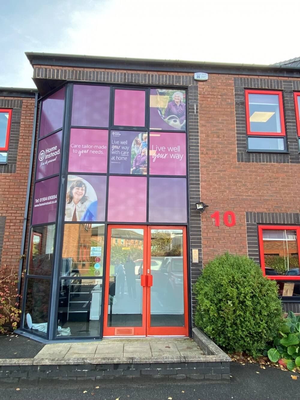 Two-story brick building with red-framed windows and a glass entrance, displaying "Home Instead" branding and signage. - Home Instead