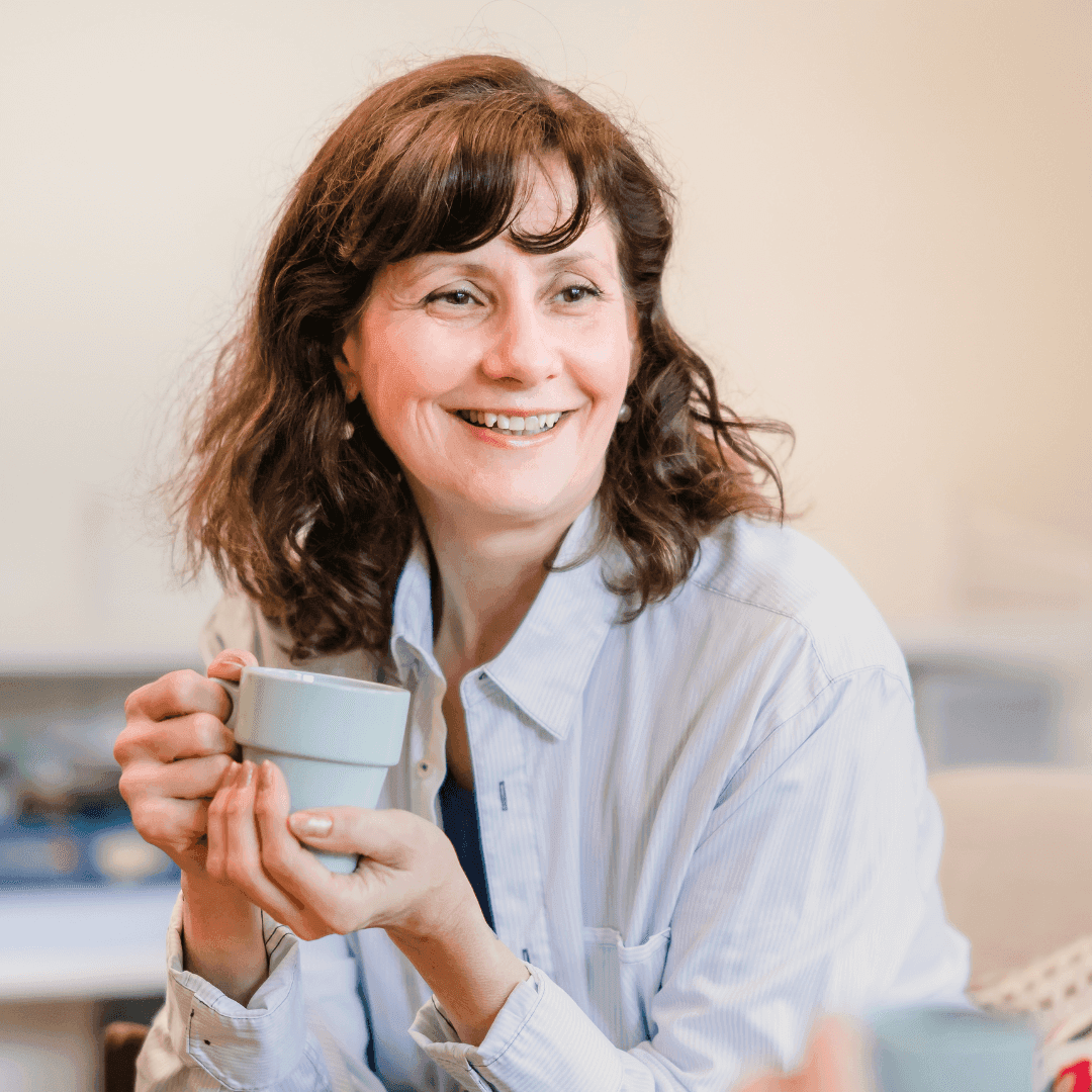 A woman with shoulder-length brown hair smiles while holding a white mug with both hands in a relaxed setting. - Home Instead