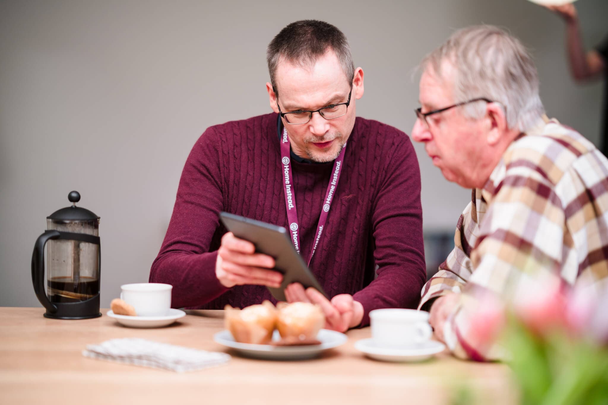 A man shows a tablet to an elderly man at a table with coffee, a French press, and pastries. - Home Instead