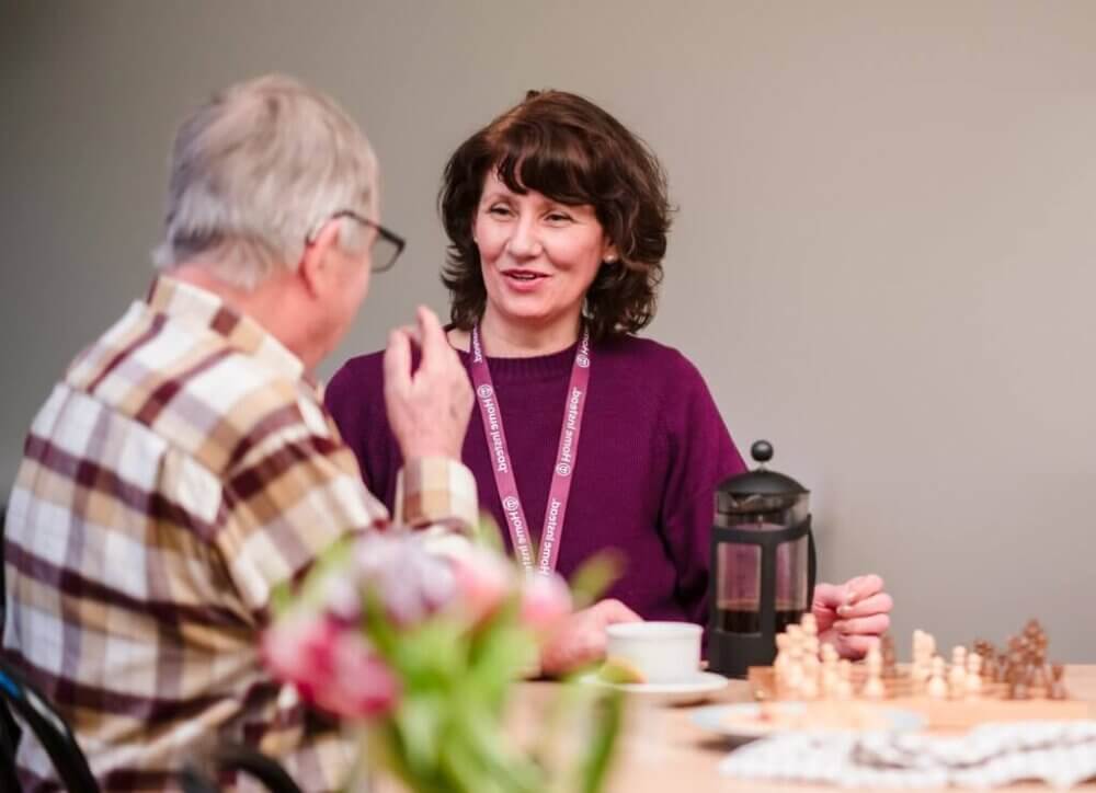 A man and a woman chatting at a table with a chess set. - Home Instead