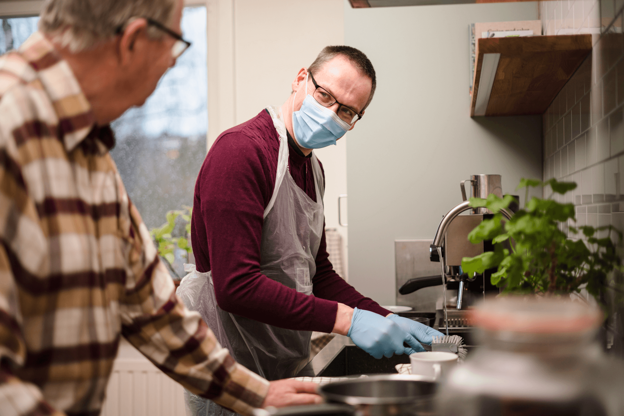 Two men cleaning dishes in kitchen; one wears a mask, gloves, and apron, while the other, in plaid shirt, watches. - Home Instead