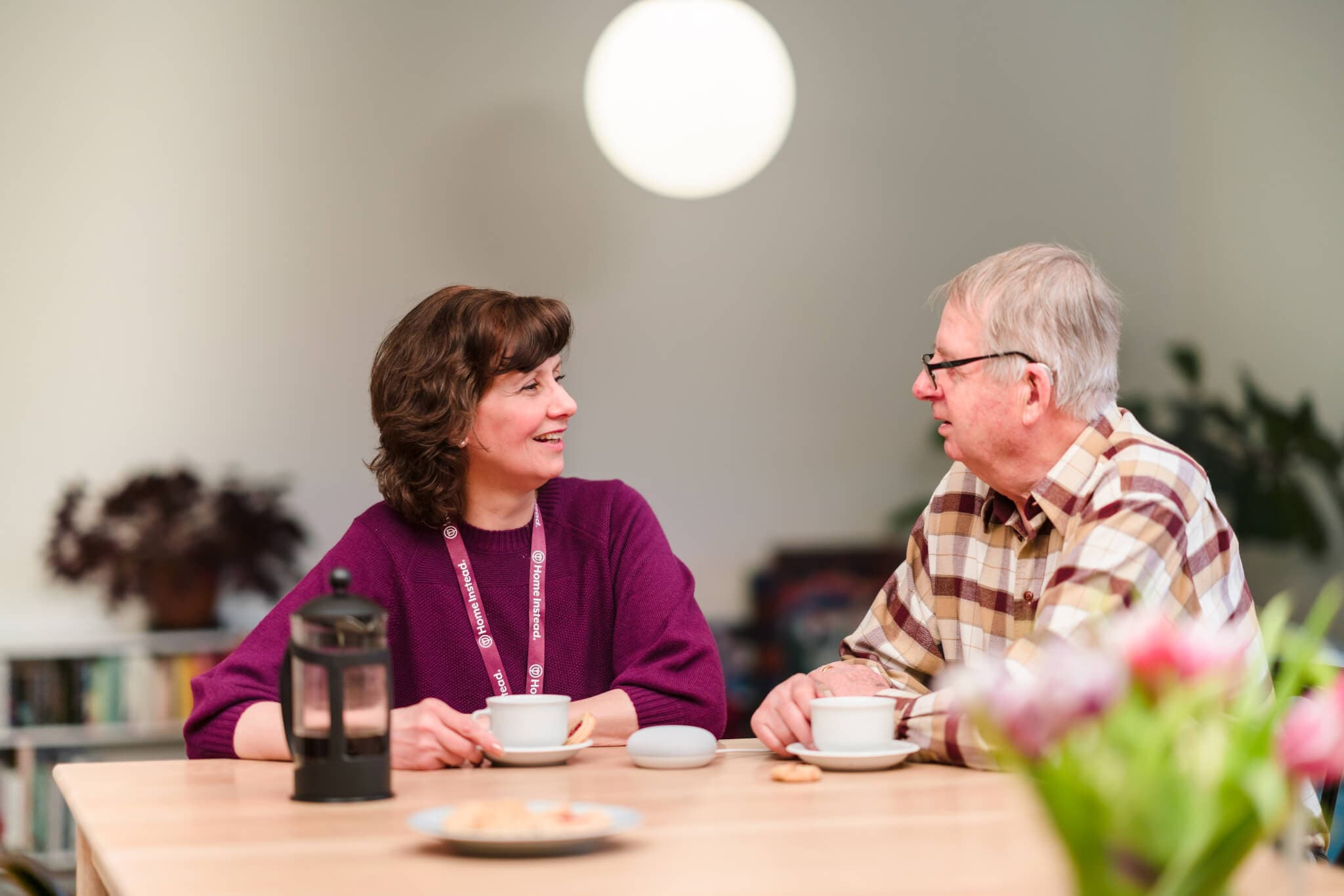 Two people are sitting at a table in a cozy setting, enjoying cups of coffee and engaging in a conversation. - Home Instead