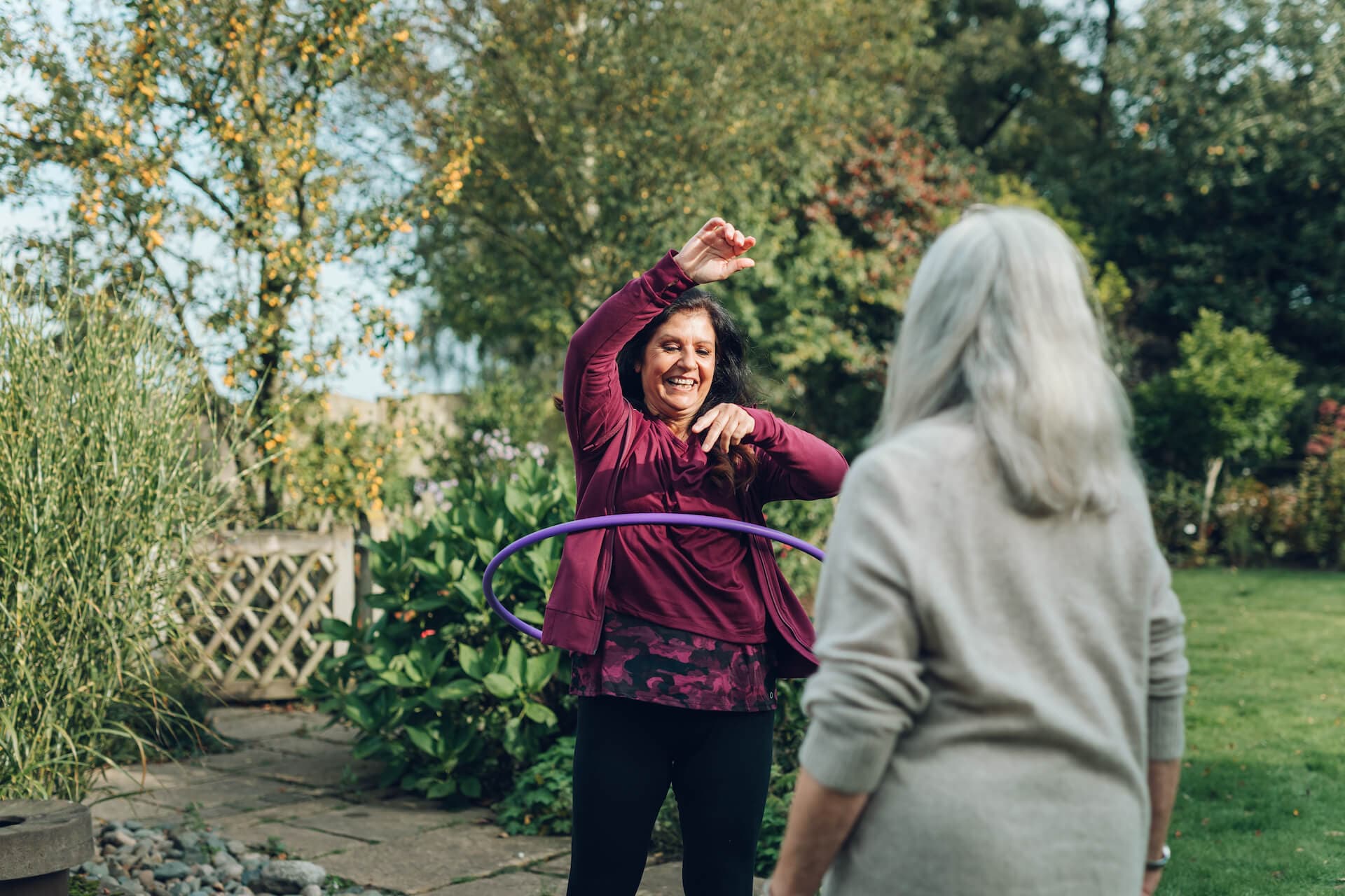 A woman hula hooping and smiling while another woman watches in a garden. - Home Instead
