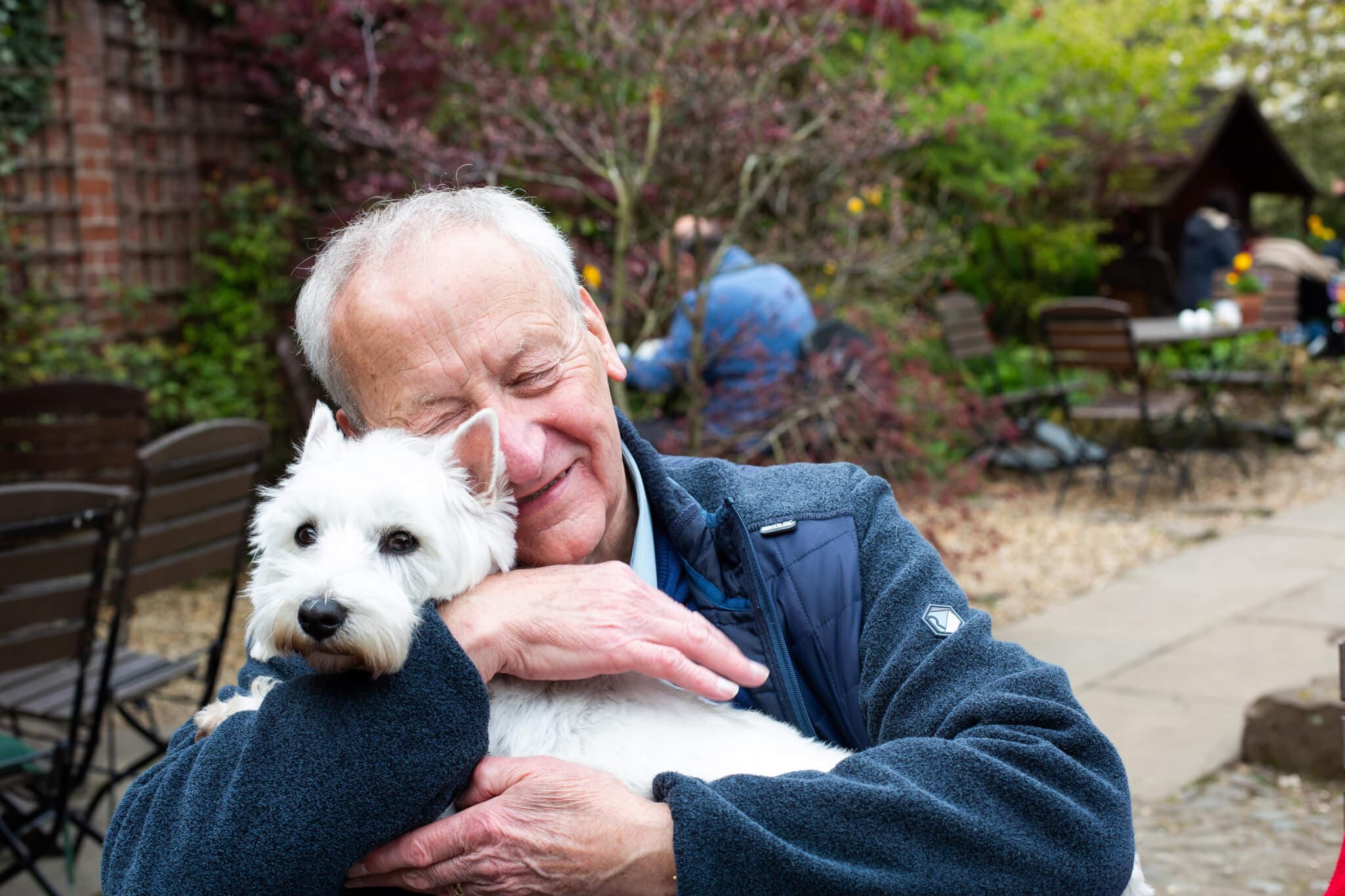 Elderly man smiling with eyes closed, hugging a white dog in an outdoor garden setting. - Home Instead