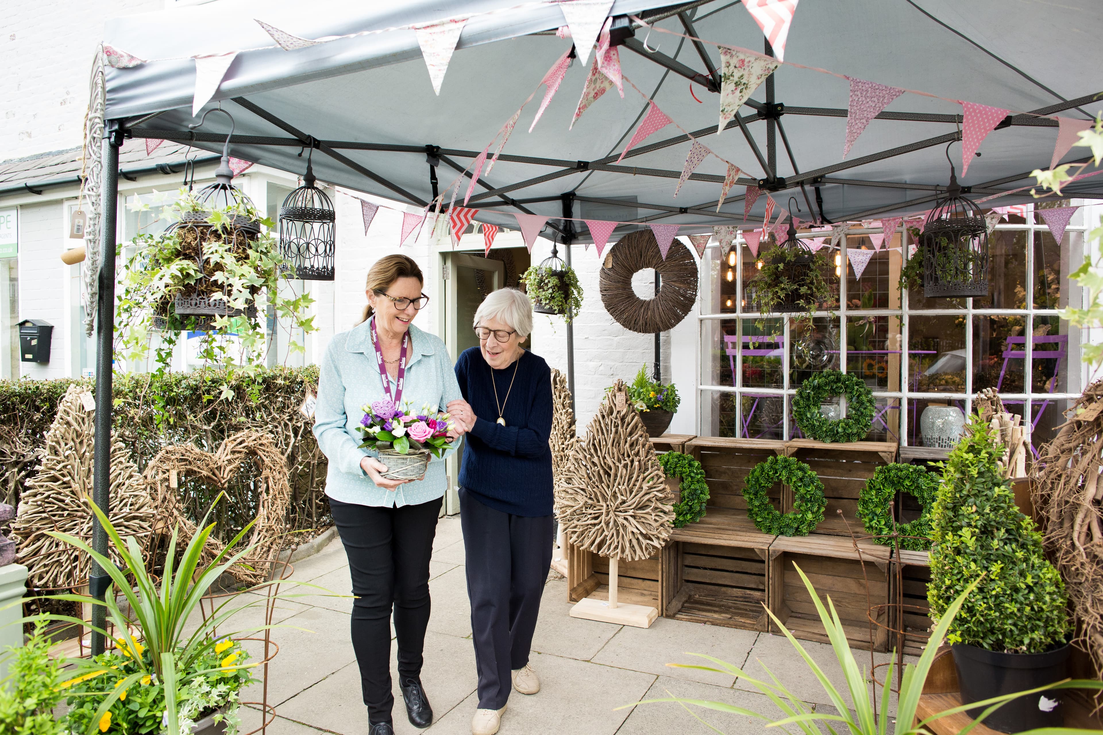 Two women, one holding a flower arrangement, smiling and walking under a decorated canopy with plants and wreaths. - Home Instead
