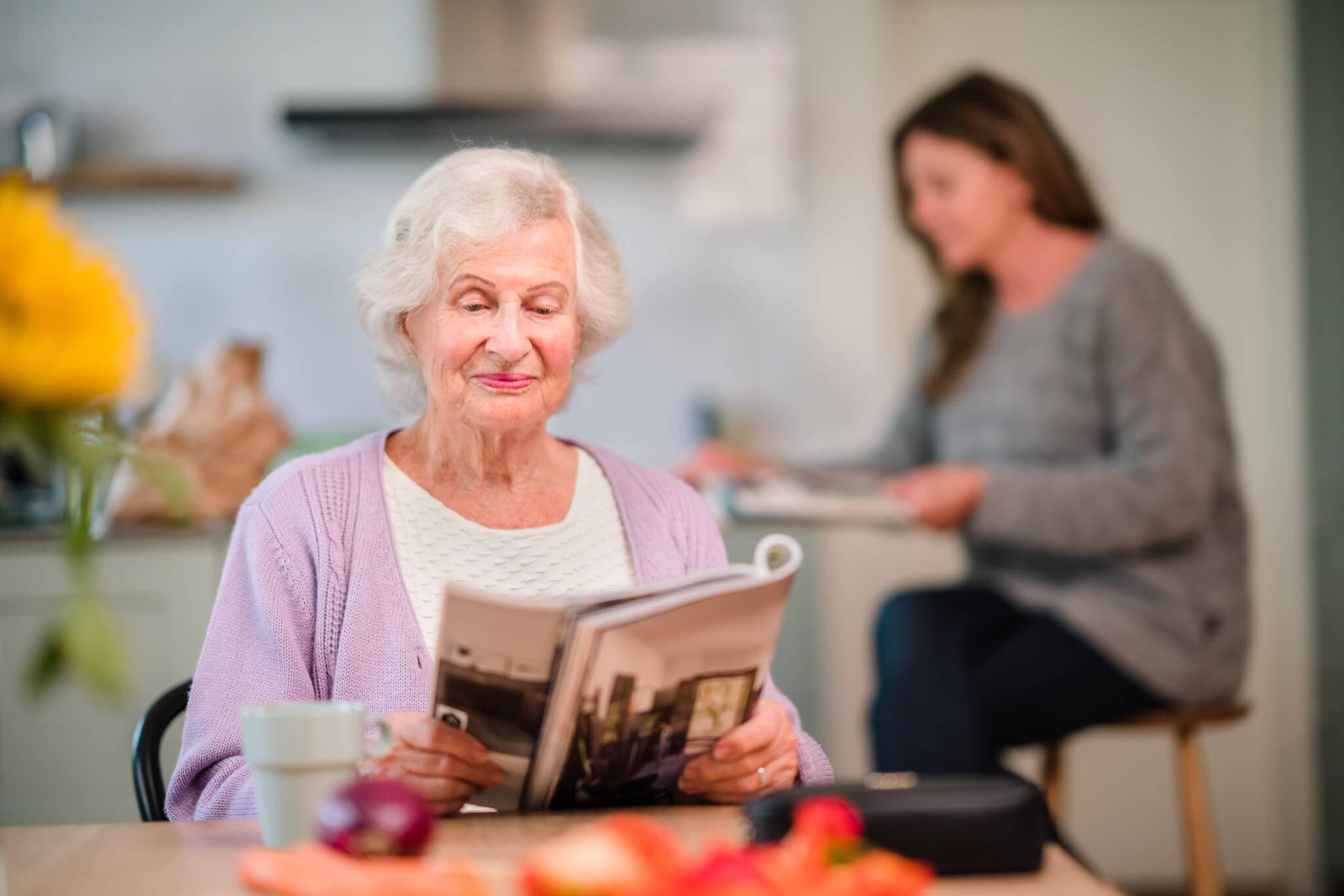 Elderly woman reading a magazine at a table with a younger woman in the background. - Home Instead