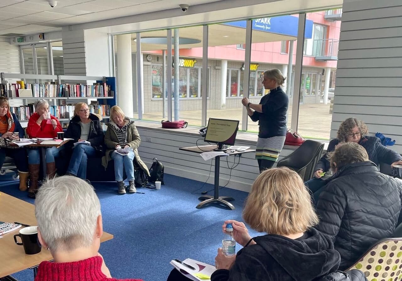 A group of people attentively watching a woman presenting near a laptop in a room with bookshelves and large windows. - Home Instead