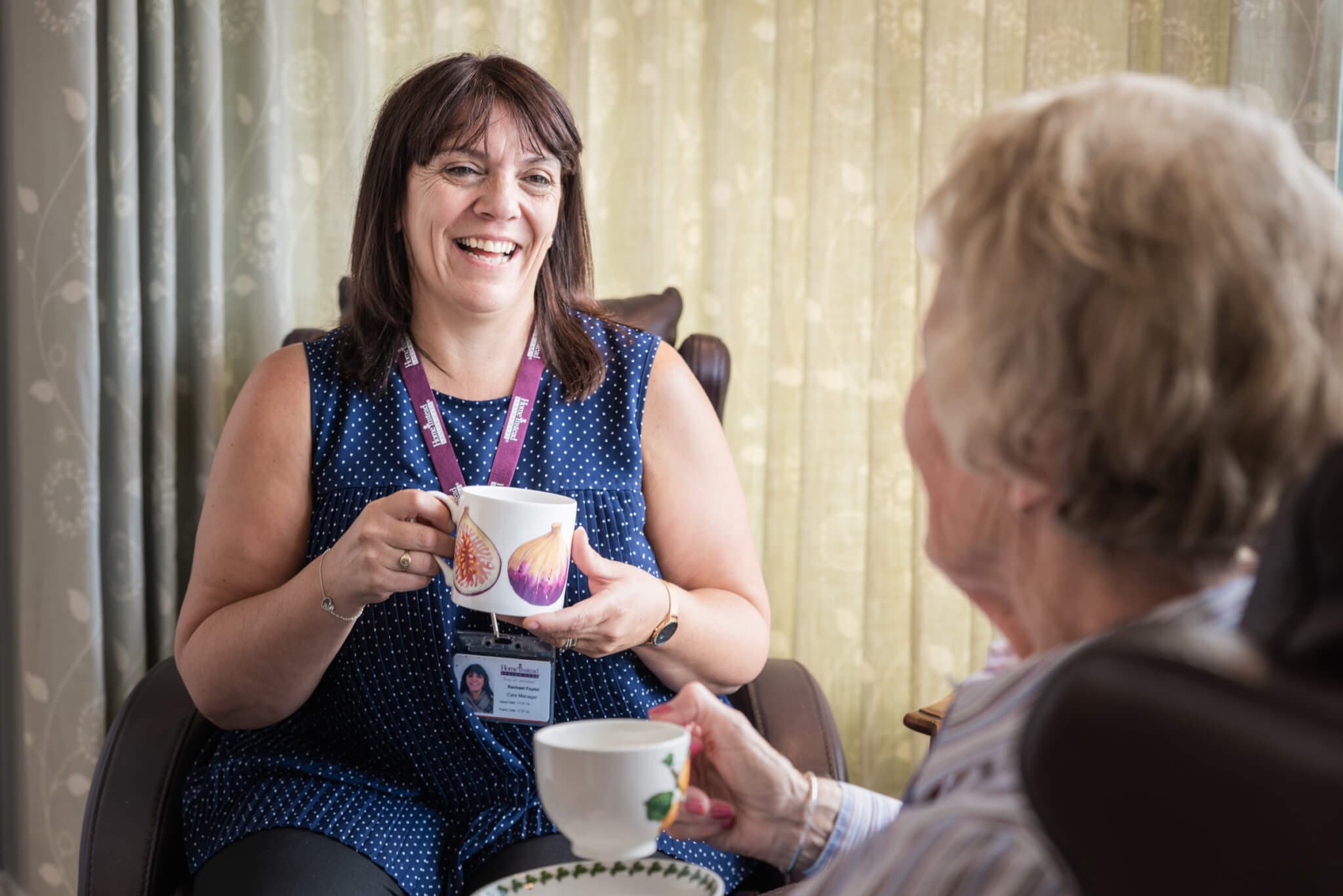 A woman smiling and holding a mug while talking with an older woman who also has a mug. - Home Instead