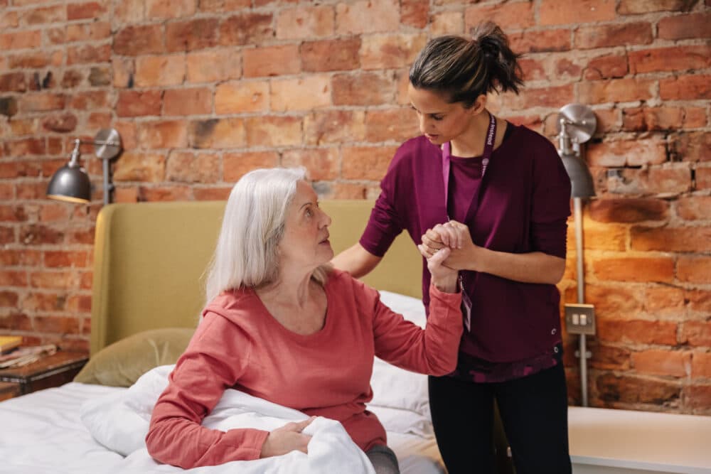 A caregiver helps an elderly woman sit up in bed in a room with exposed brick walls. - Home Instead