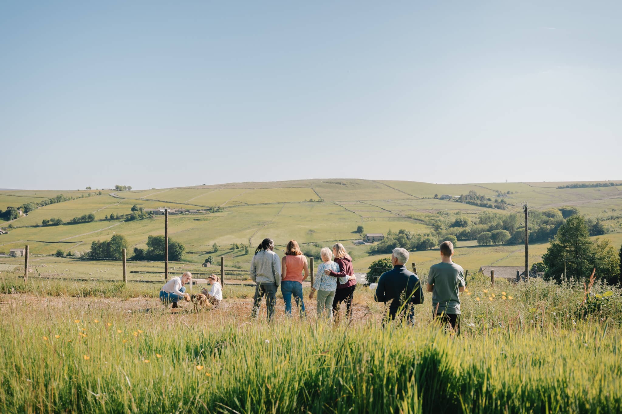 A group of people stand in a grassy field with hills and clear blue sky in the background. - Home Instead