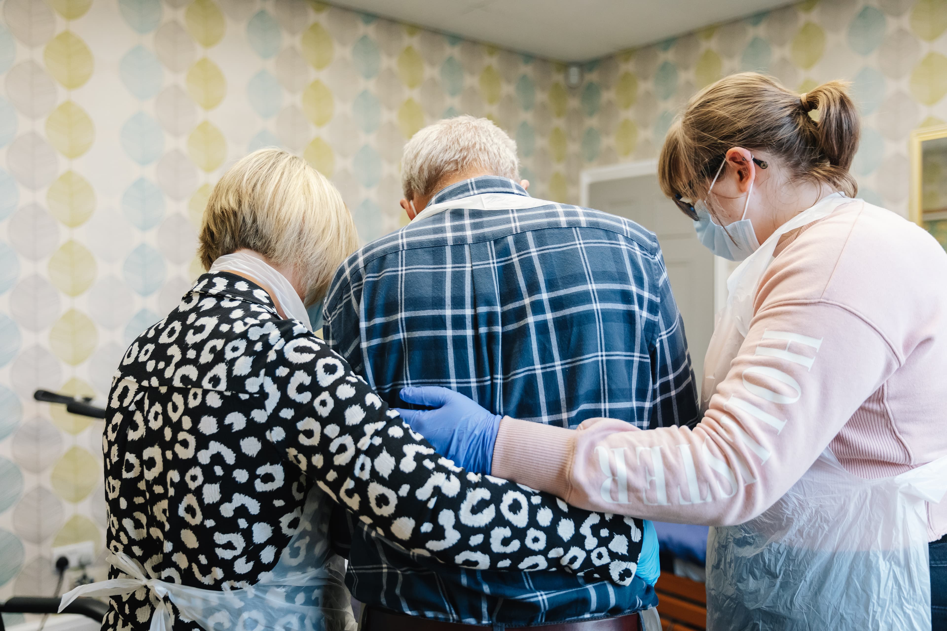 Three people standing together; two women on either side support an elderly man in a plaid shirt in the center. - Home Instead