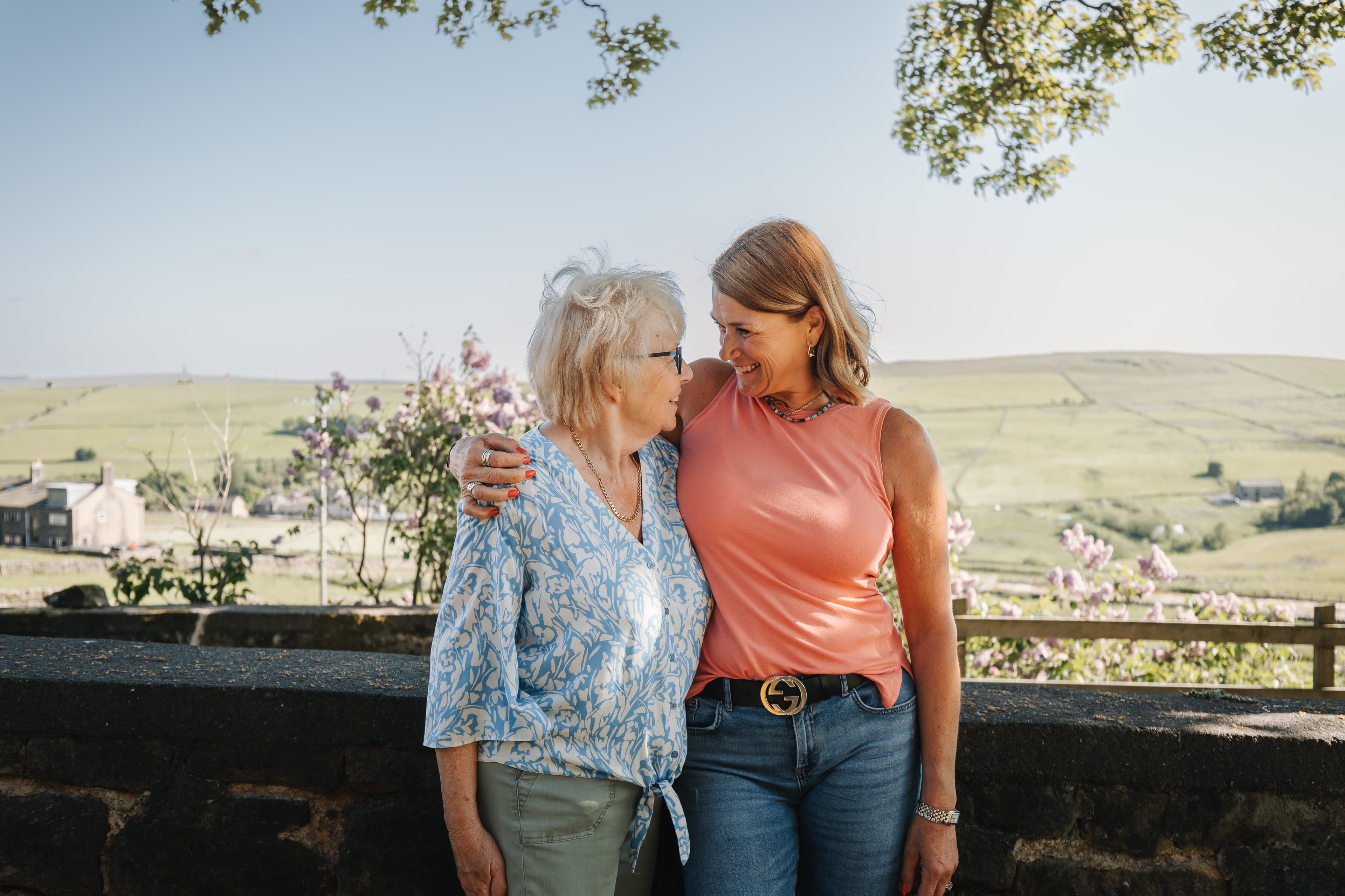 Two women, one elderly and one younger, share a warm embrace while standing in a scenic countryside setting. - Home Instead