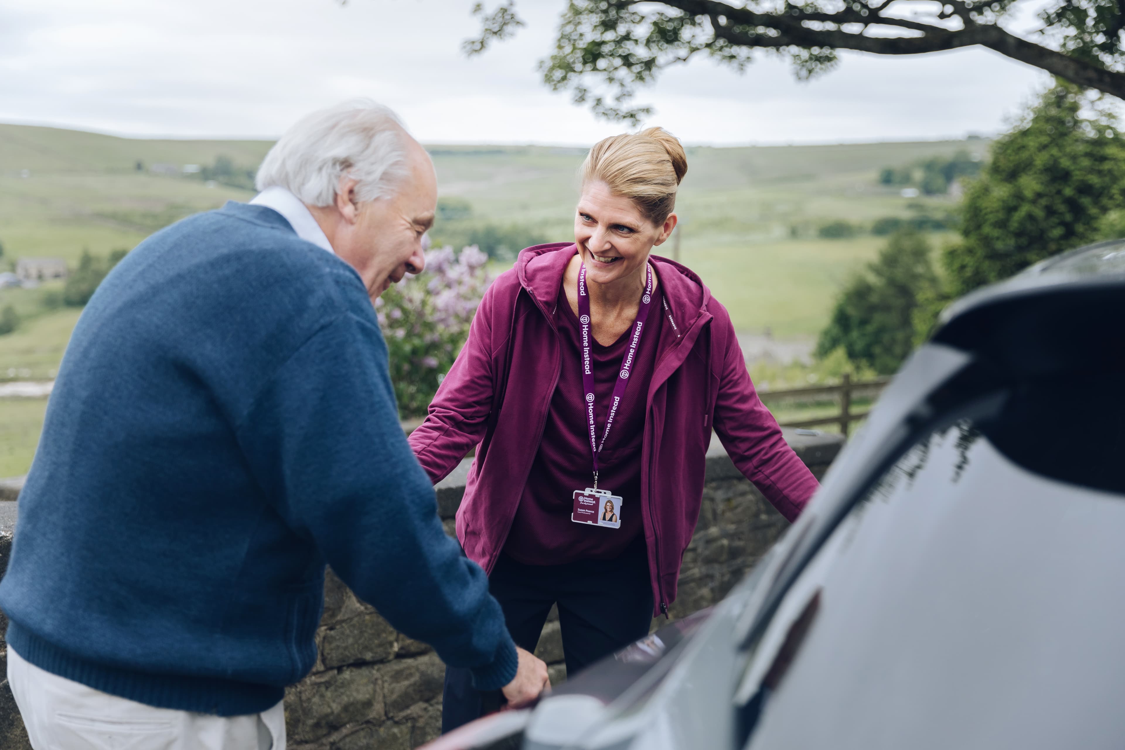 A caregiver helps an elderly man near a car in a rural setting with trees and hills in the background. - Home Instead