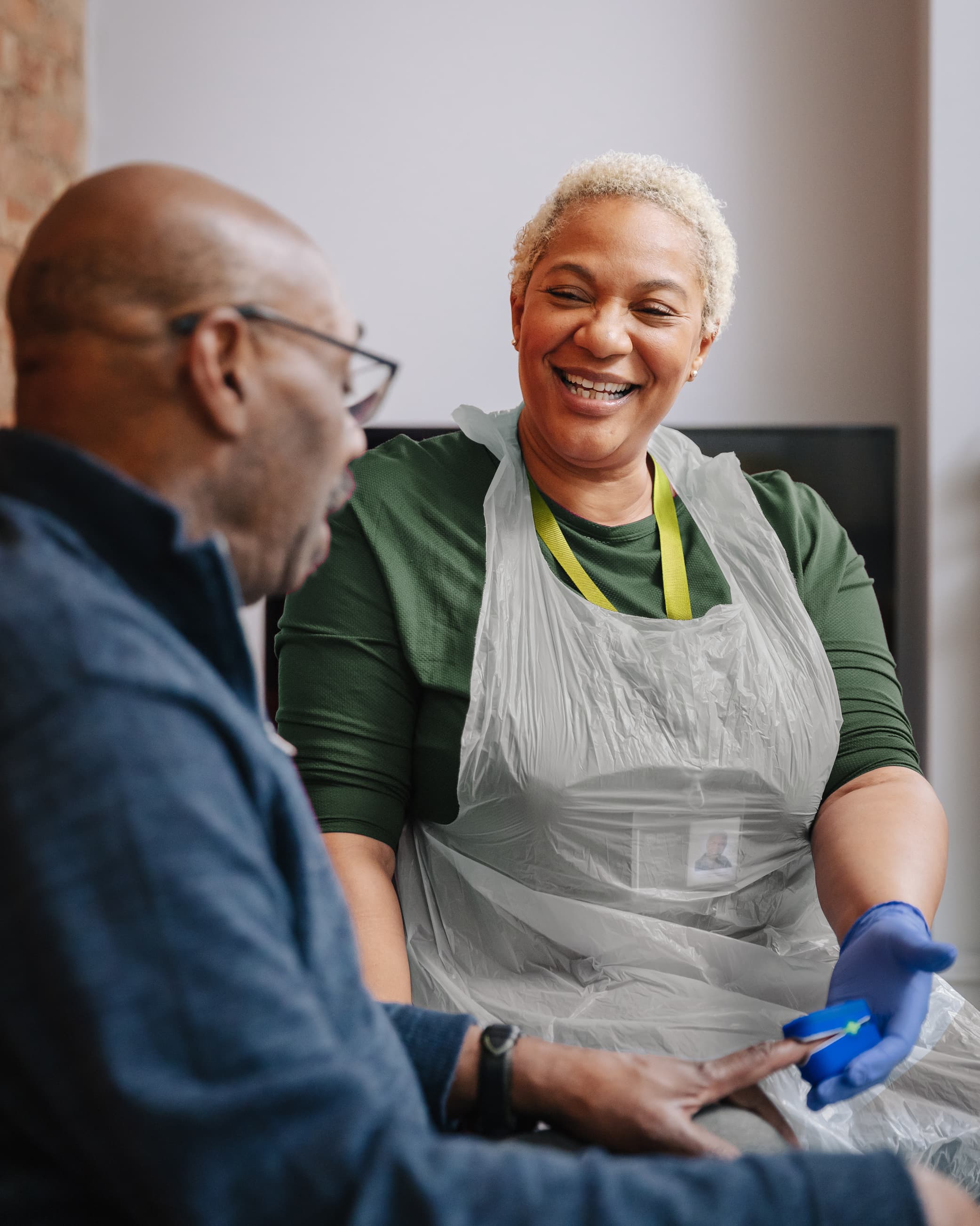 A smiling caregiver wearing gloves helps an older man, holding his hand during a health check. - Home Instead