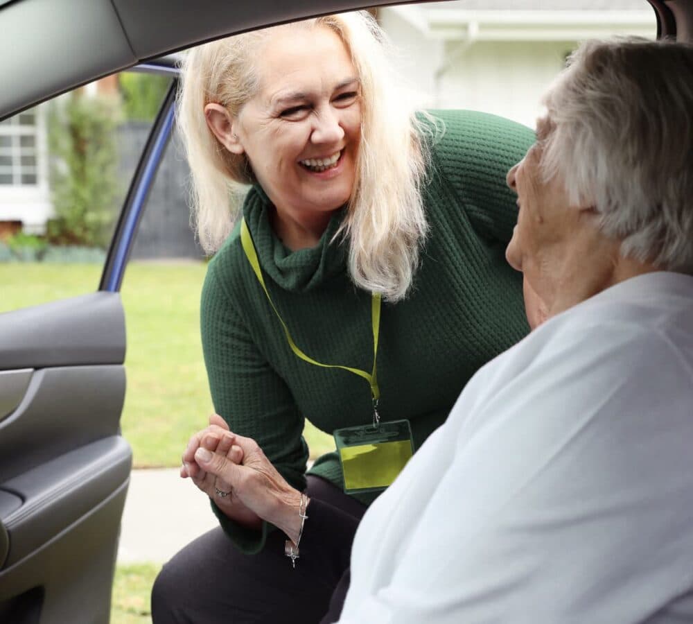 Smiling woman helps an older adult seated in a car, both appear happy and engaged outside a house. - Home Instead