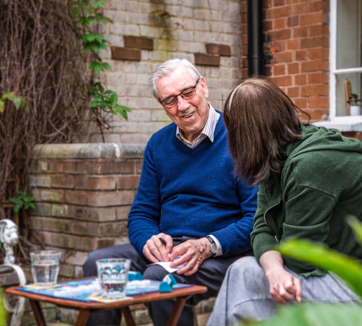 An elderly man and a woman sit outside, smiling and talking by a small table with drinks and a board game. - Home Instead