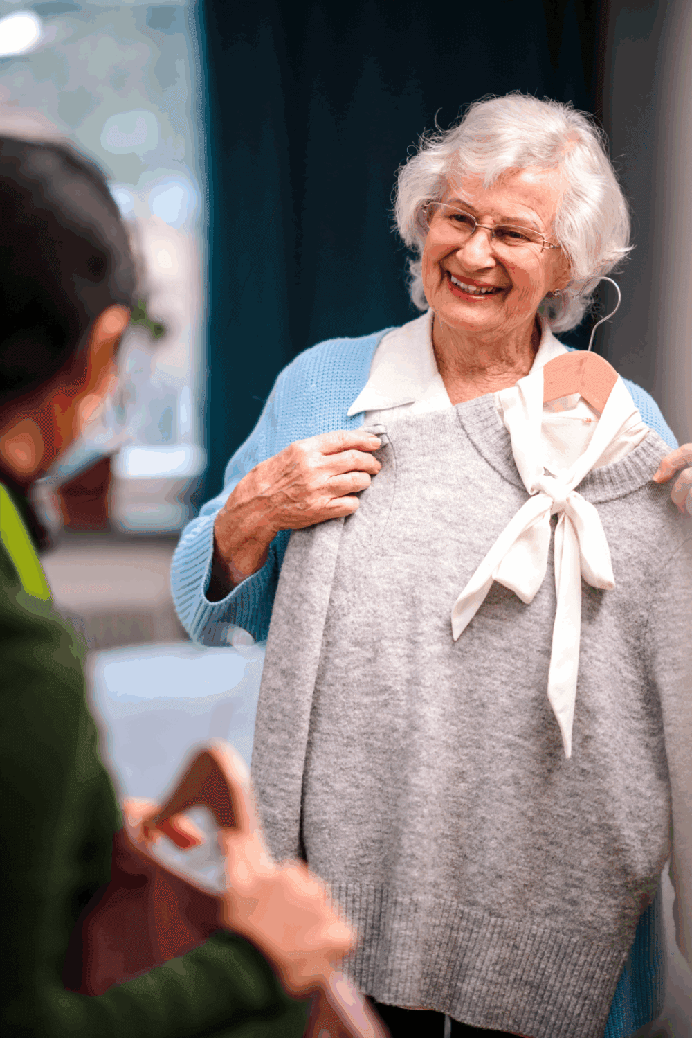 Smiling elderly woman holds up a gray sweater on a hanger while talking to another person. - Home Instead